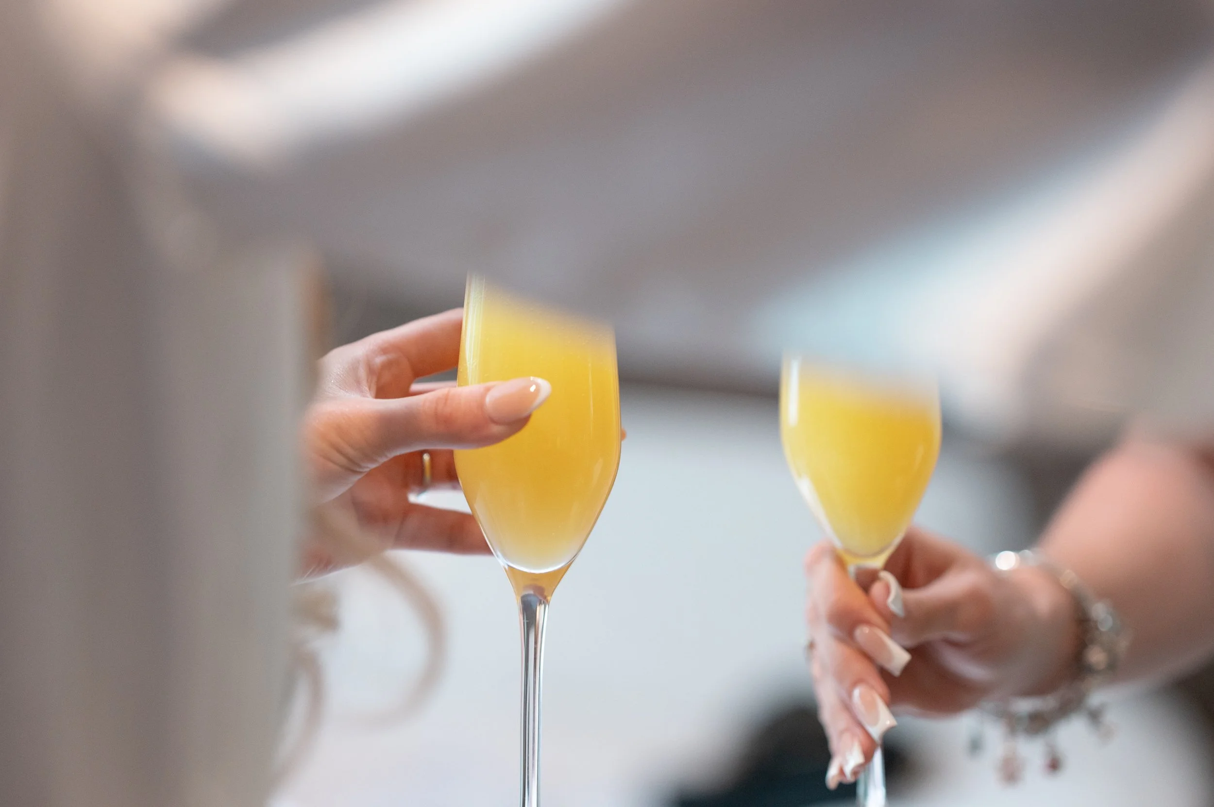 Two hands holding champagne flutes filled with yellow-orange beverage, likely at a celebration or toast.
