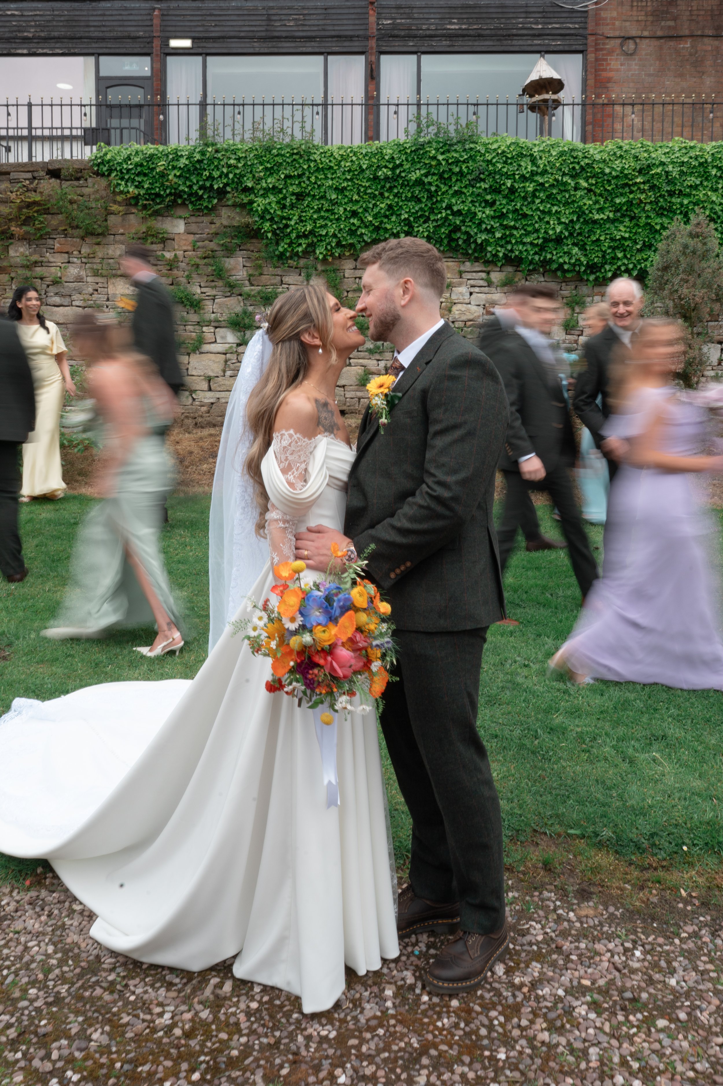 Bride and groom standing closely, smiling, holding hands, outside on grassy area during wedding, with wedding guests blurred walking by in background, and a brick wall and ivy-covered fence behind them.