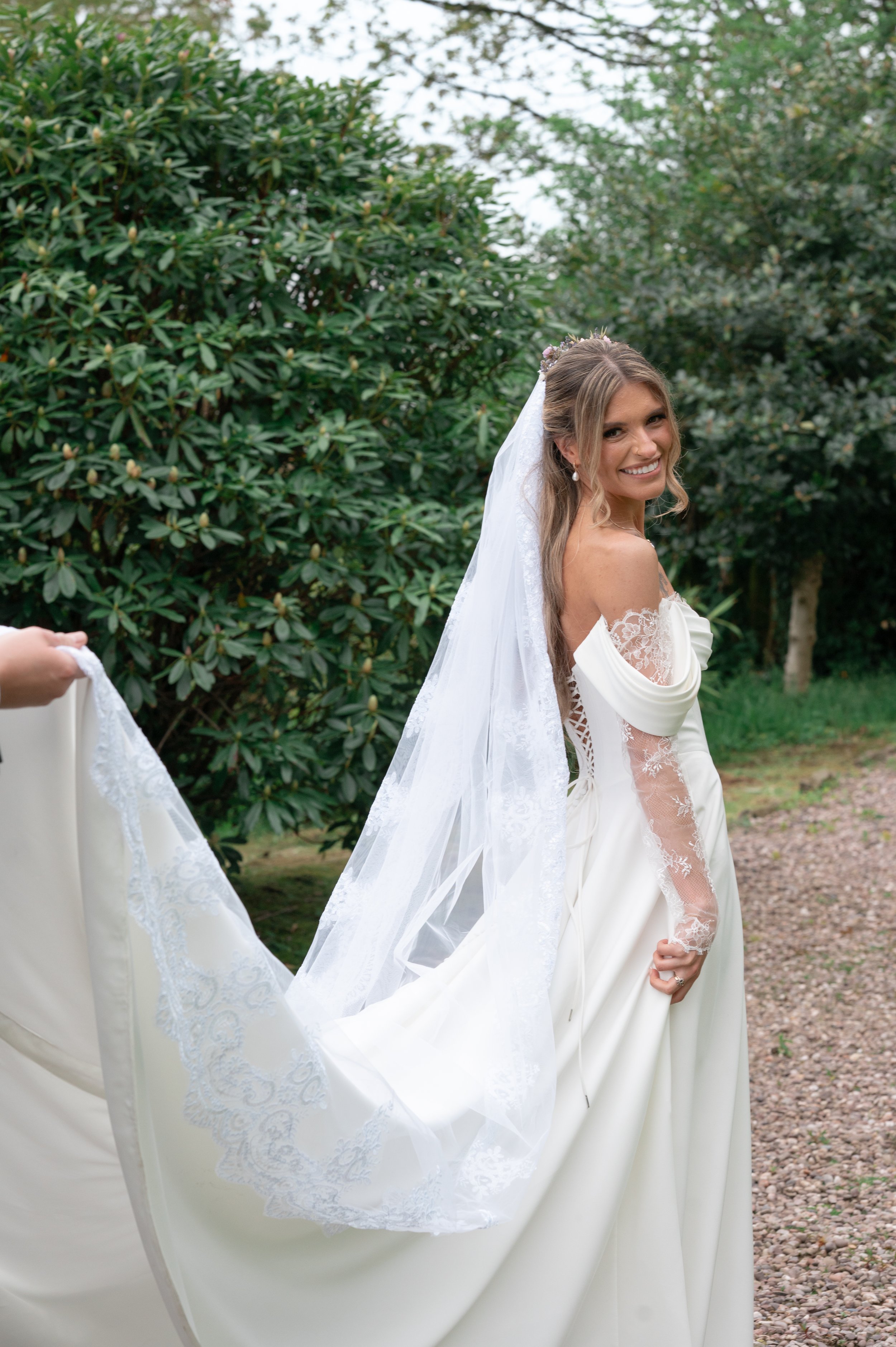 A smiling bride in a white wedding dress with lace details, standing outdoors with green bushes and trees in the background.