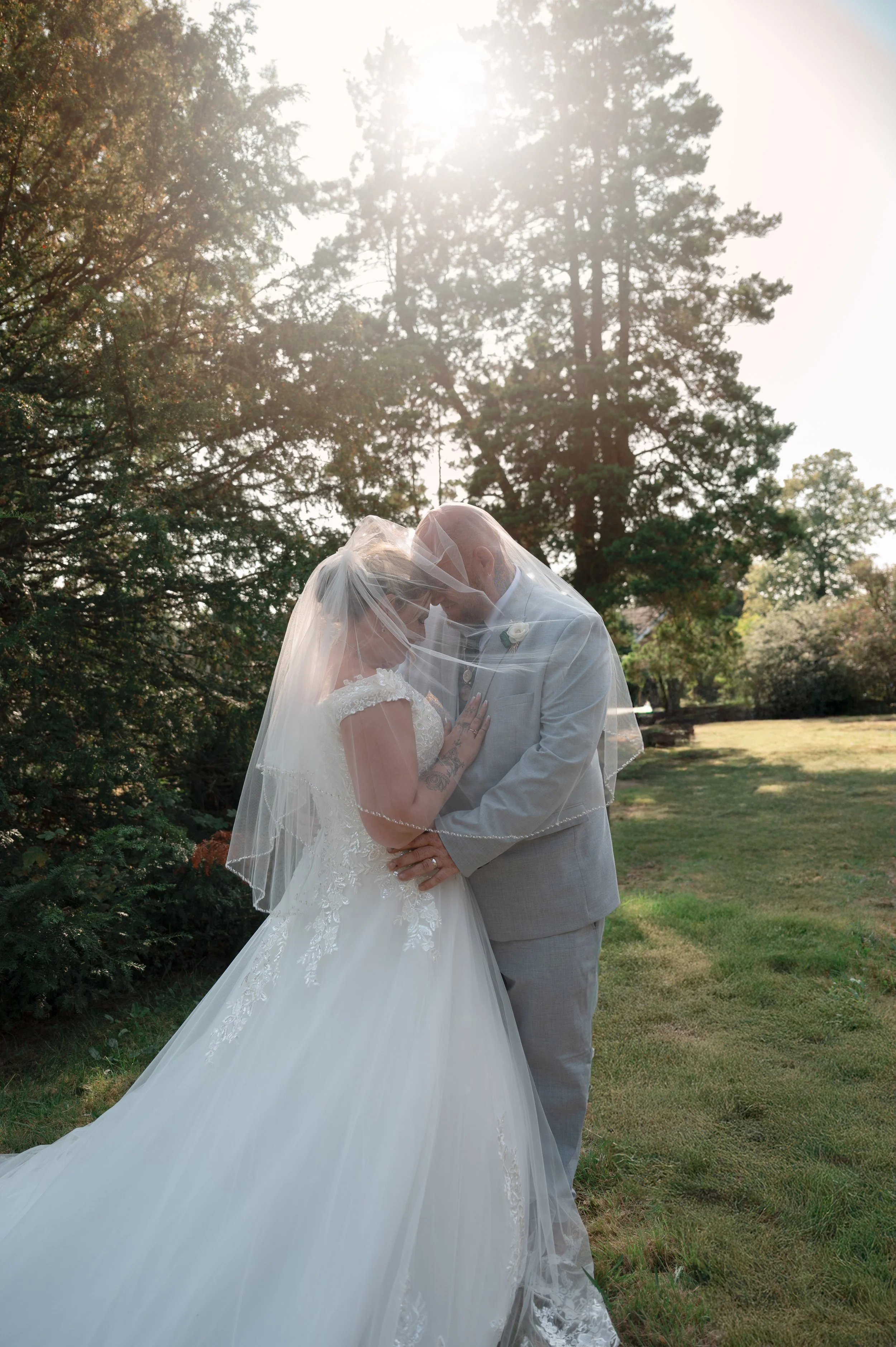 A bride and groom dressed in wedding attire, standing close together outdoors on a grassy area with trees in the background, under bright sunlight.