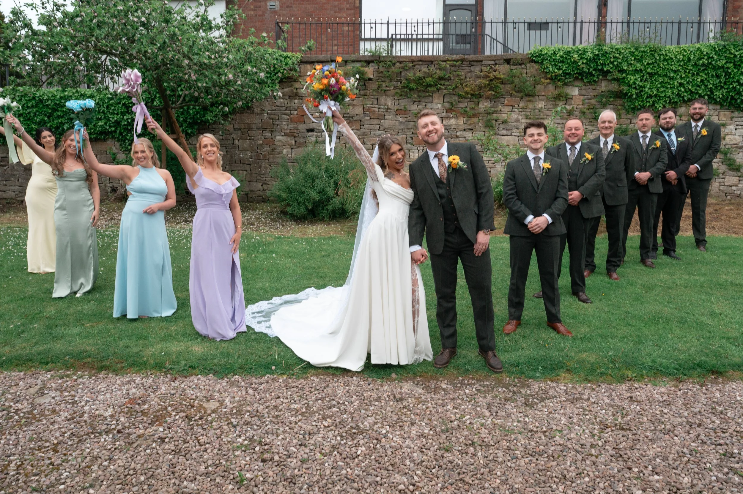 People at a wedding celebration, including a bride in a white gown holding a bouquet and a groom in a suit. Bridesmaids in pastel dresses and groomsmen in suits with yellow boutonnieres pose outdoors in front of a stone wall and greenery.