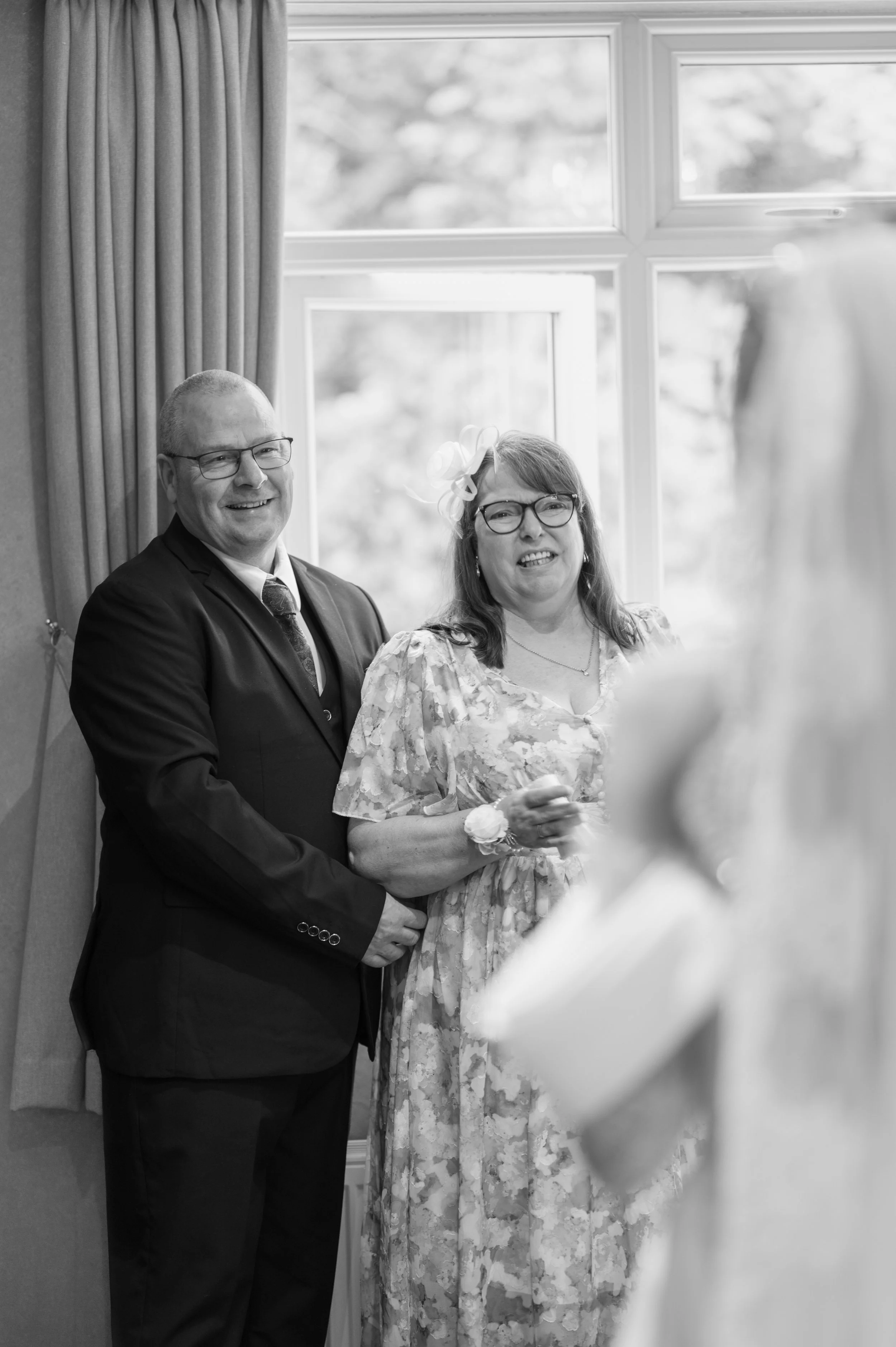A smiling couple, a man in a suit and a woman in a floral dress and glasses, standing together indoors near a window with curtains, during a social event or celebration.