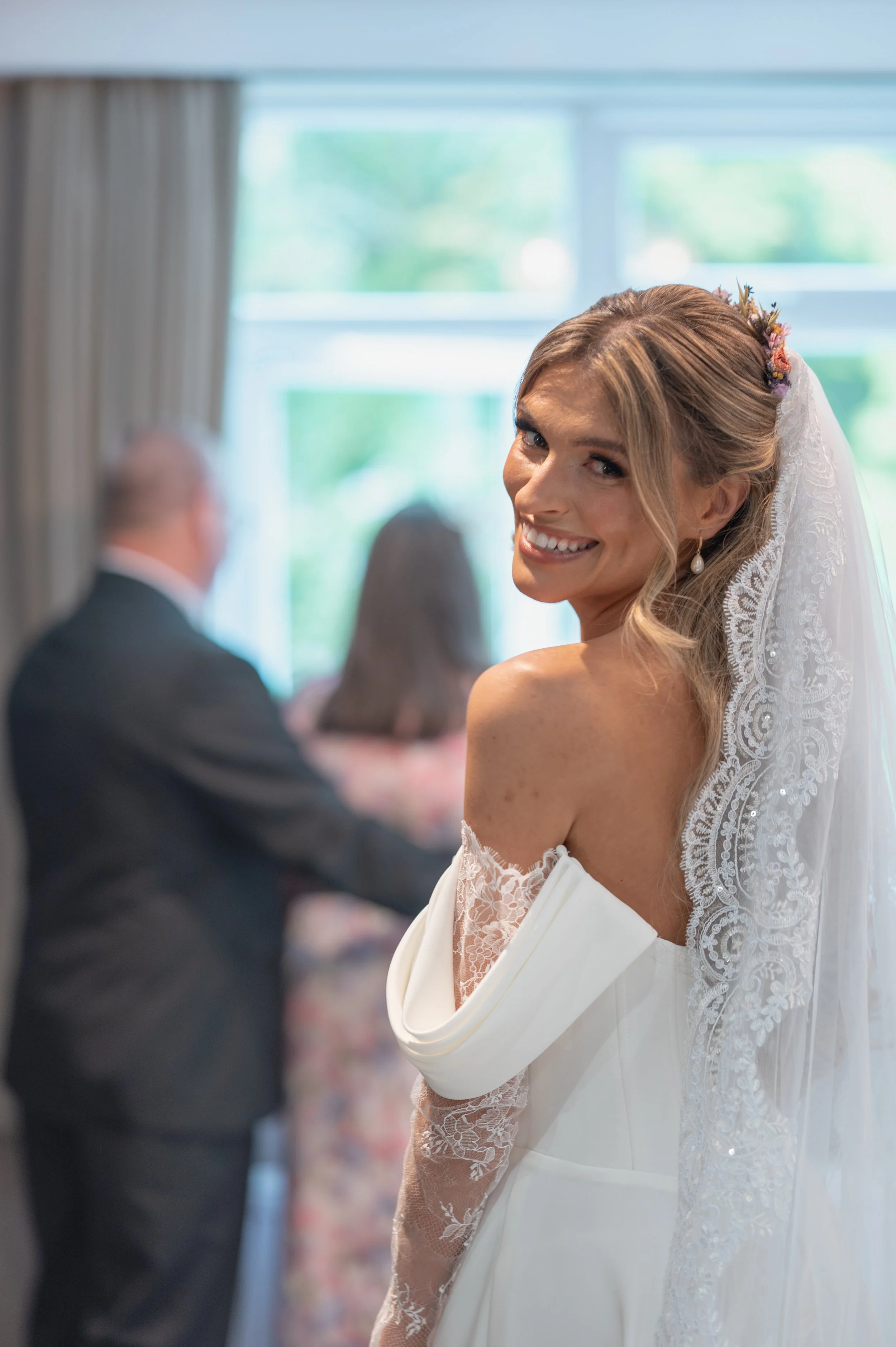 A smiling bride with light brown hair styled in loose waves, wearing a white wedding dress with lace sleeves, pearl earrings, and a floral hairpiece, standing at her wedding ceremony with blurred guests in the background.