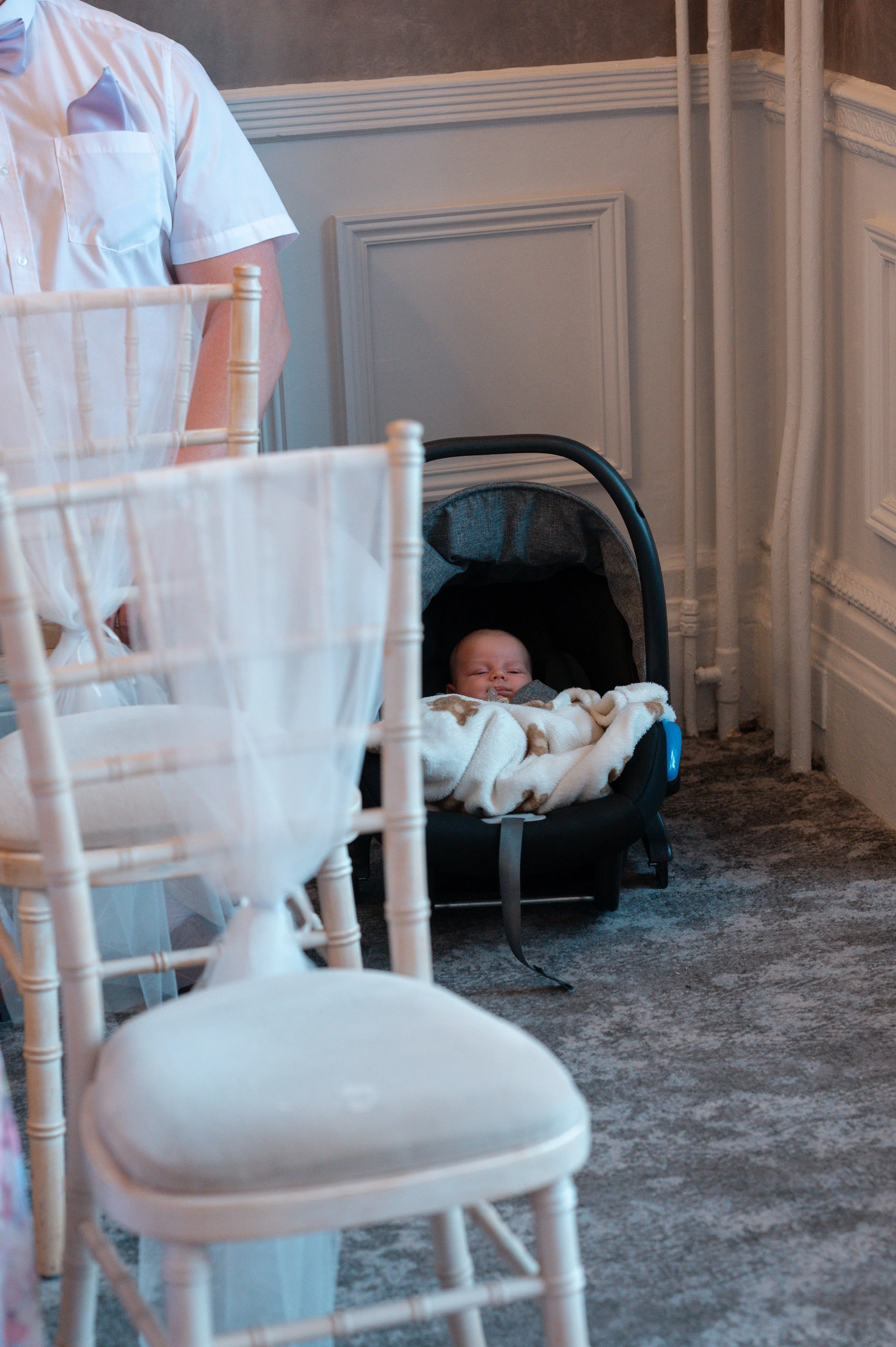 A baby asleep in a car seat covered with a blanket, with part of a person visible standing nearby, in a decorated room with white chairs and wainscoting.