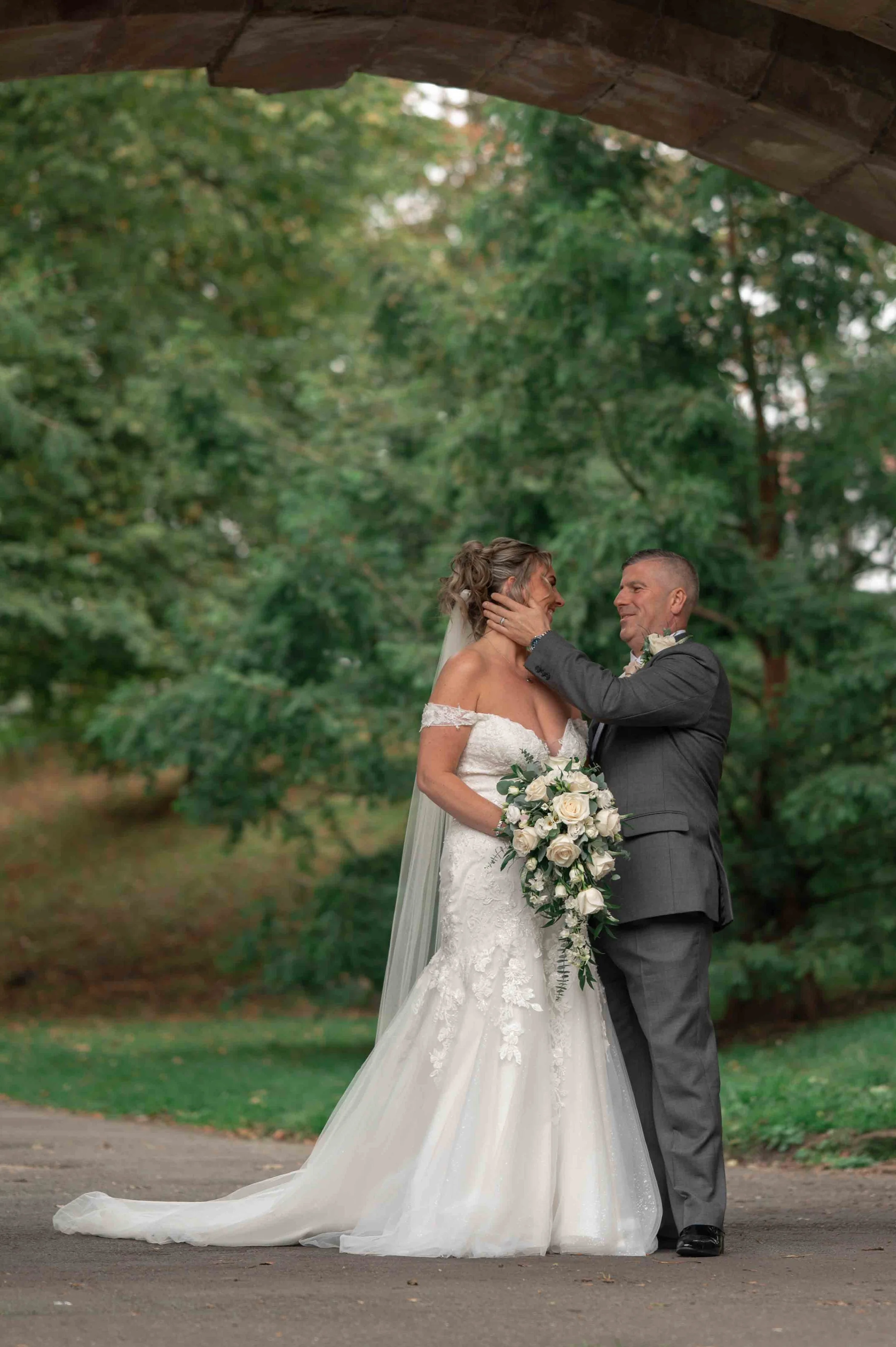 A bride and groom stand close together beneath a brick arch, sharing an intimate moment outdoors, with surrounding green trees in the background.