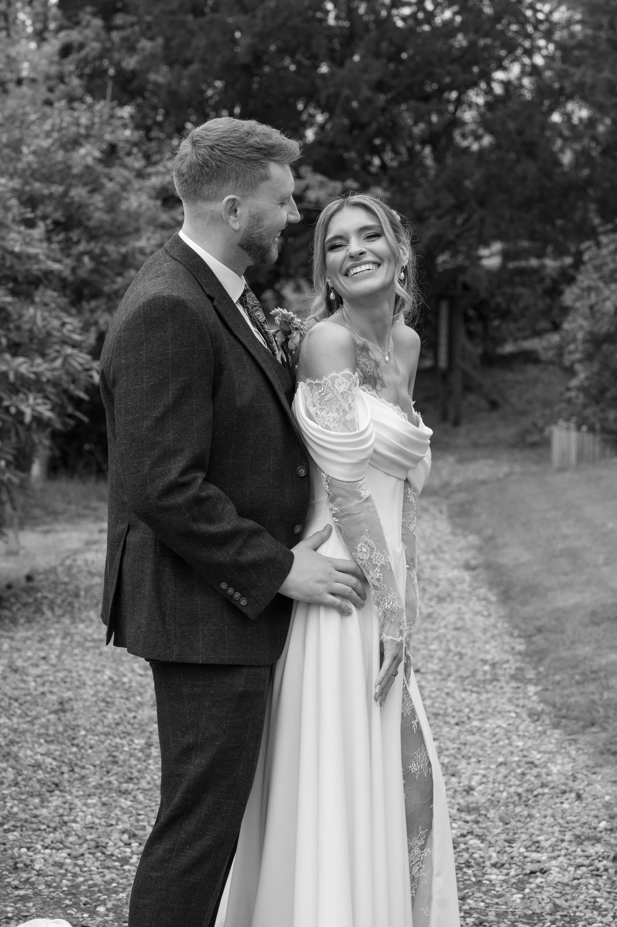 A black and white photo of a happy couple on their wedding day outdoors. The groom wears a dark suit with a patterned tie, and the bride is in a white wedding dress with lace details on the sleeves. They are smiling and looking at each other, standin