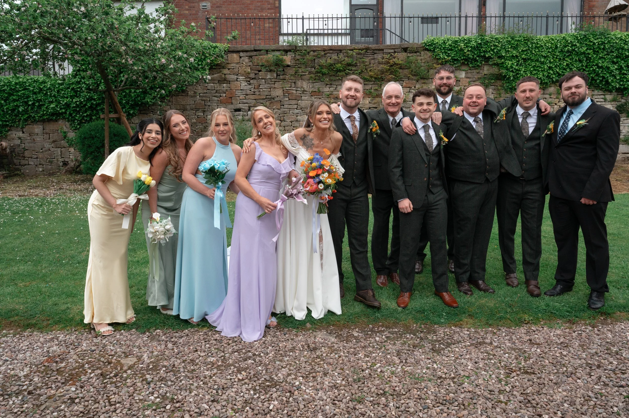 Group of people at a wedding, including women in pastel dresses and men in black suits, standing outdoors in front of a brick wall with greenery.