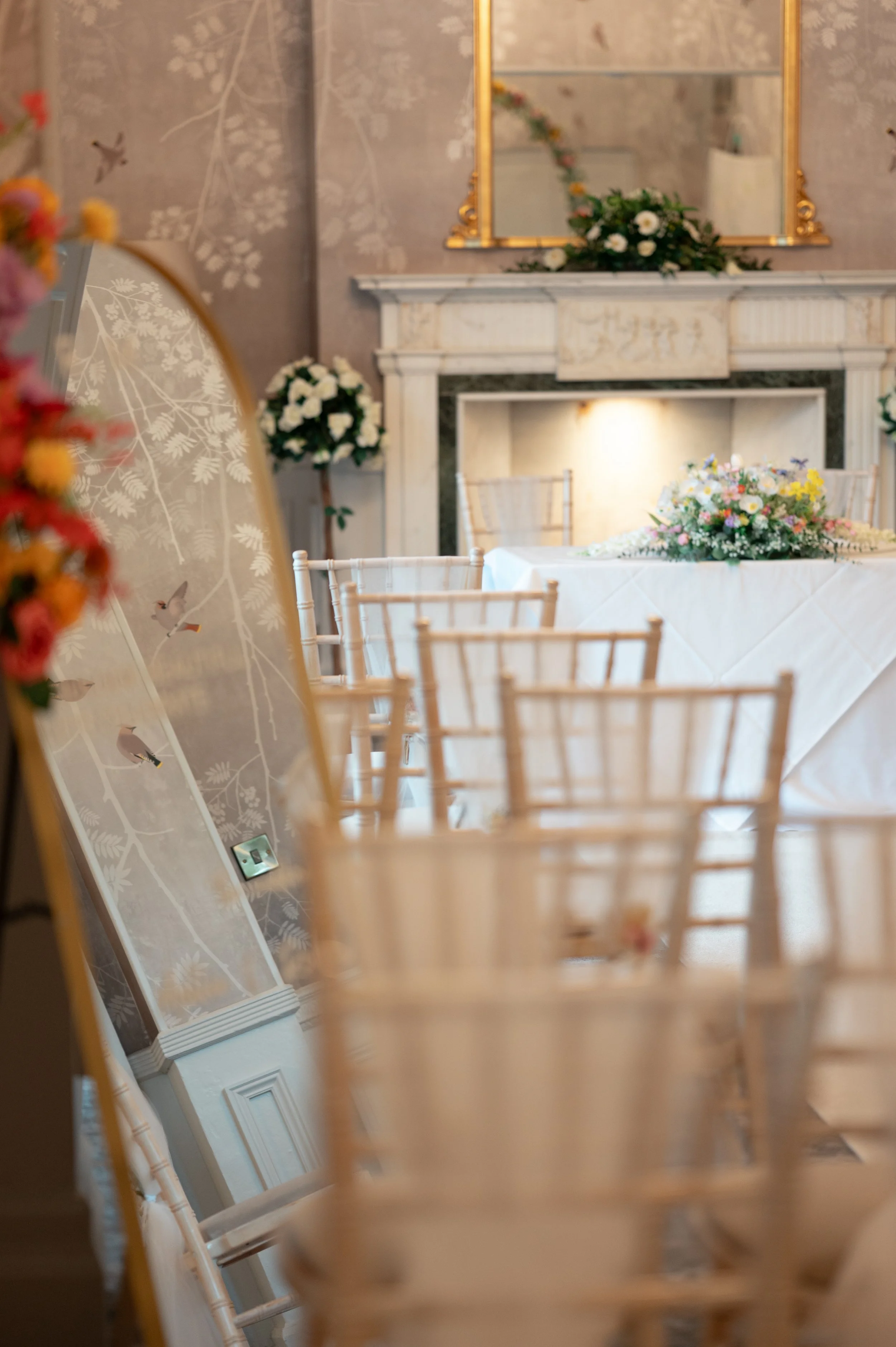 A decorated interior space with a floral centerpiece on a table, a large ornate mirror, and chairs. The room has a fireplace with floral arrangements and patterned wallpaper with birds.