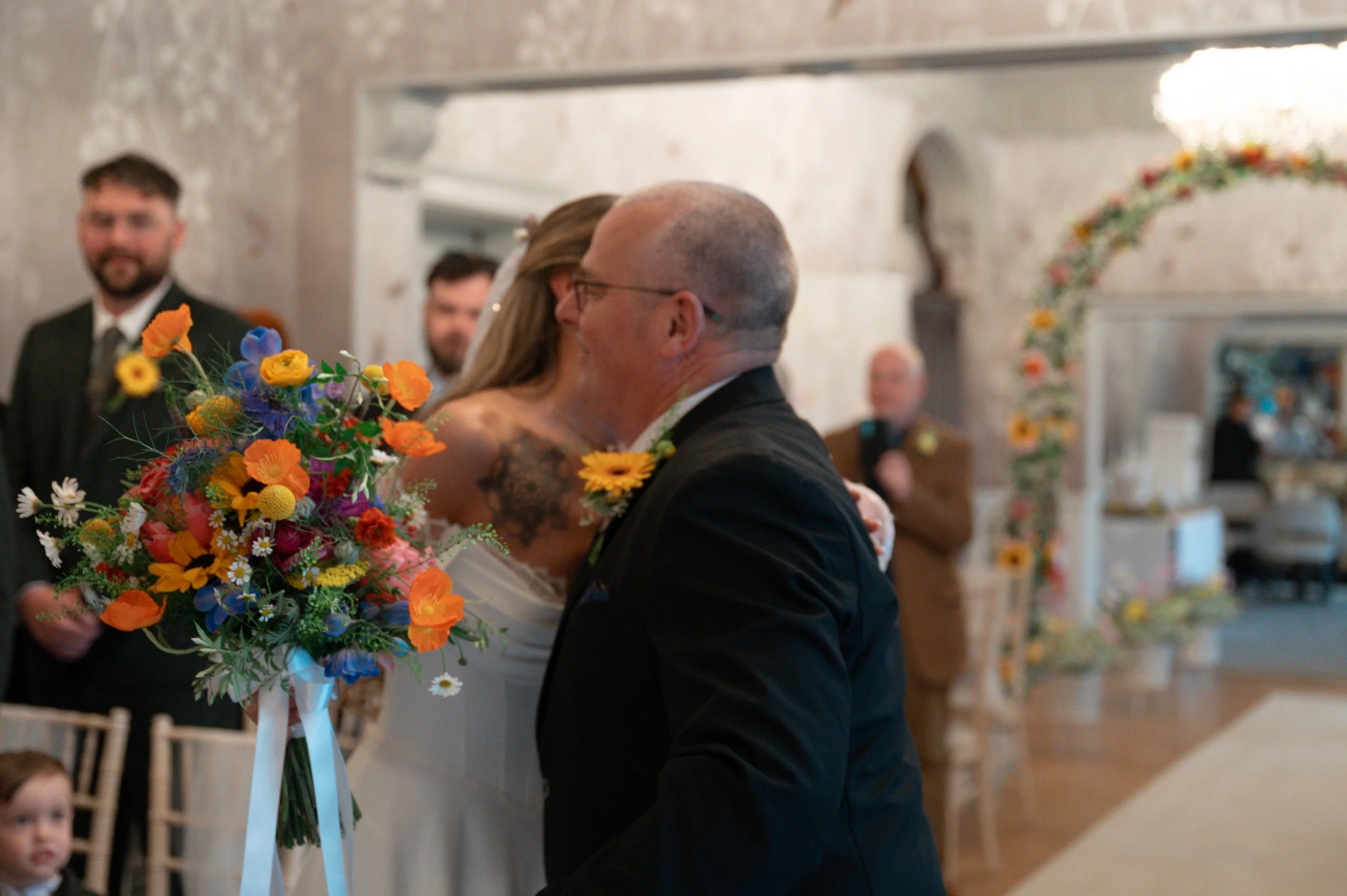 A bride and groom sharing a kiss during a wedding ceremony, with a bouquet of colorful flowers in the foreground and guests in the background, inside a decorated venue.