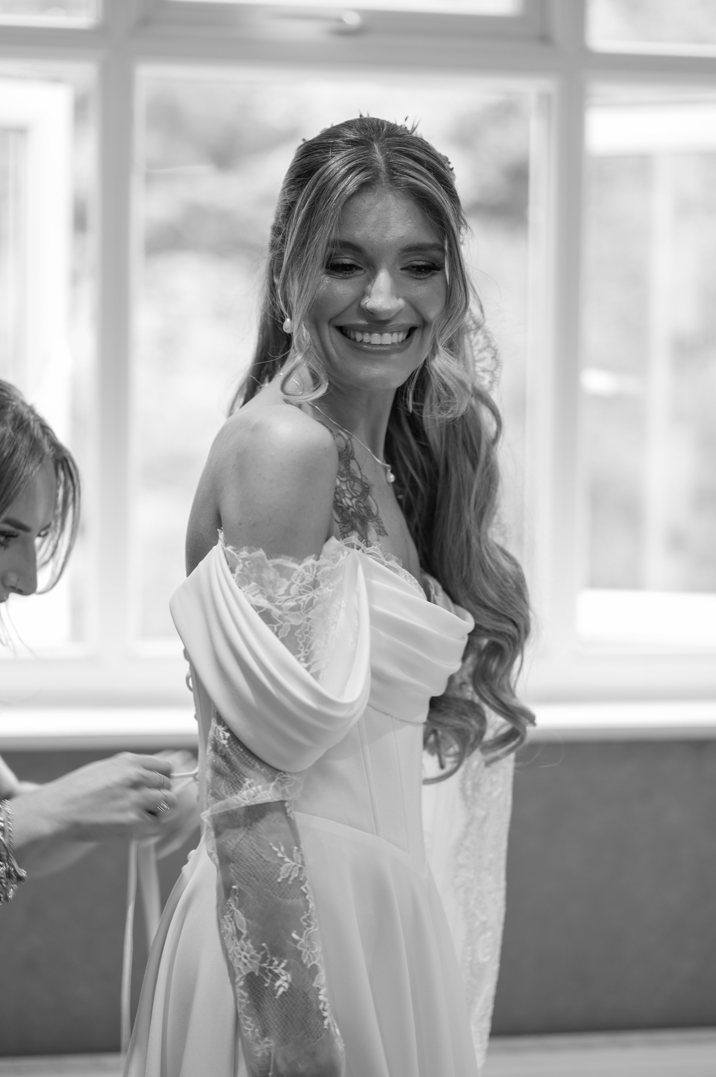 A woman in a wedding dress smiling while a person behind her helps with her dress in a room with large windows.