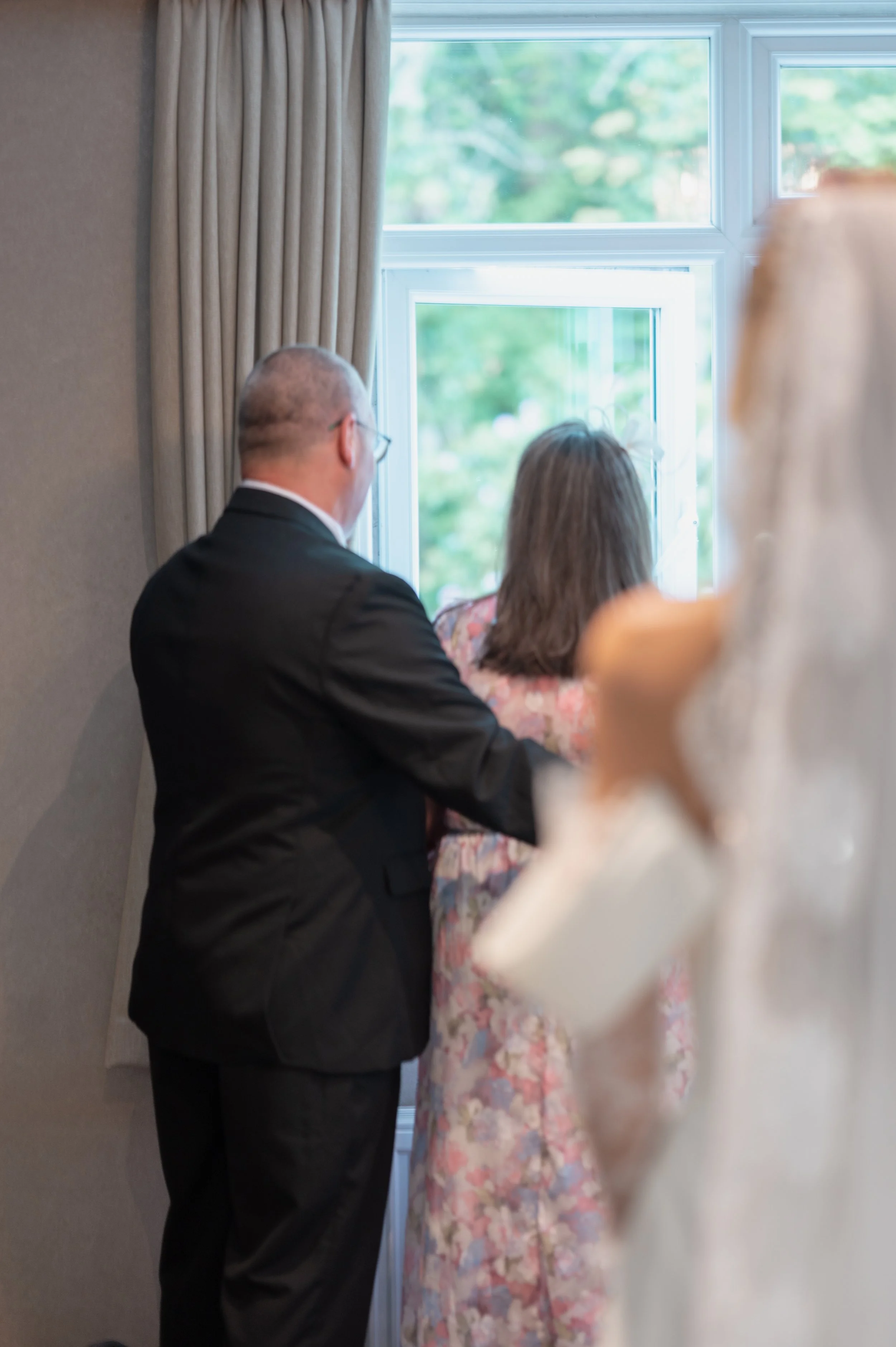 A man in a black suit with glasses and a woman in a floral dress are standing together by a window, looking outside. The scene is inside a room with beige curtains and trees visible through the window.