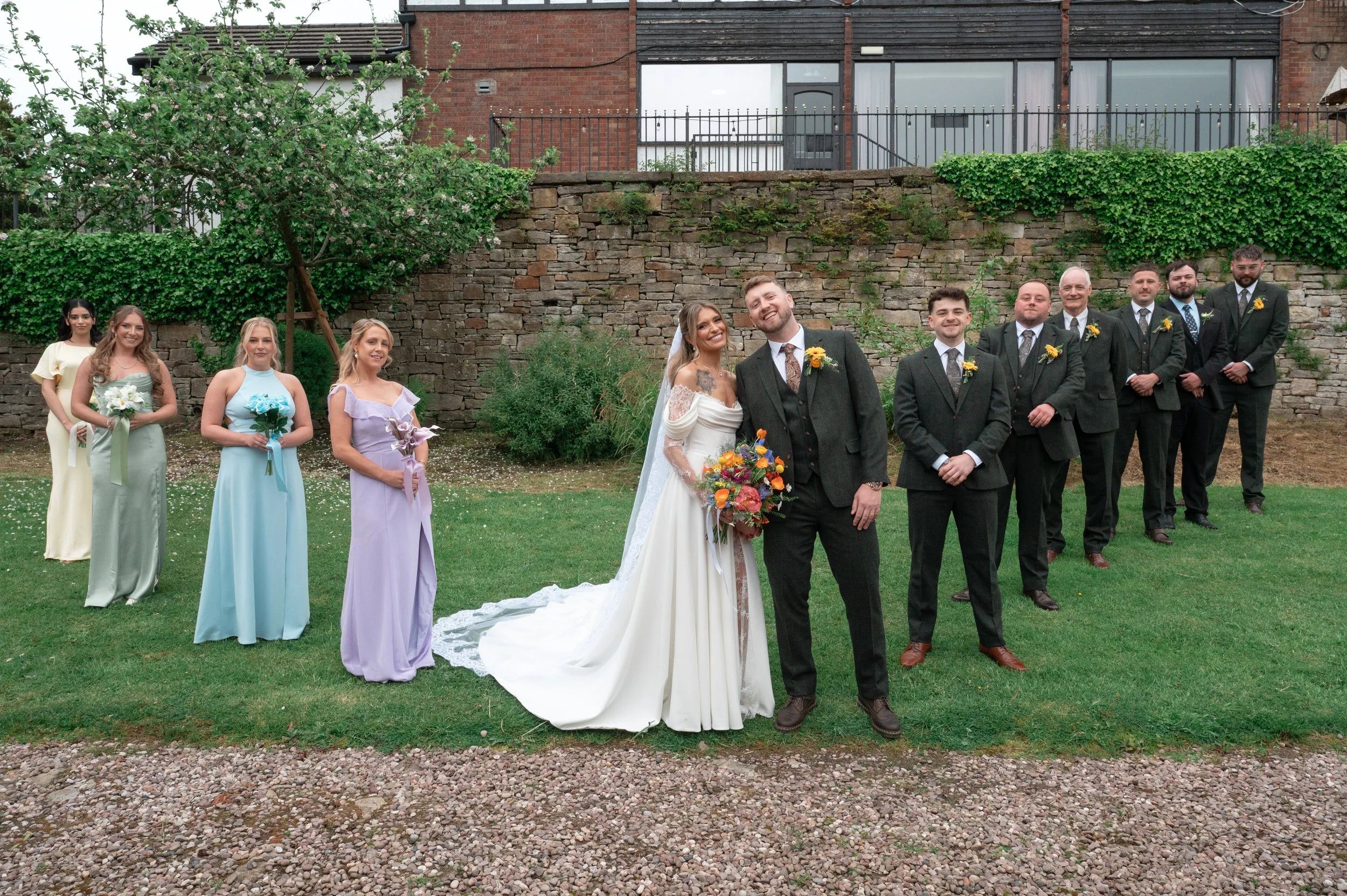 A group of ten people, including a bride and groom, standing outdoors on a grassy area with a brick wall and building in the background. The bride wears a white wedding dress with a train, and the groom wears a dark suit with a patterned tie. Six men