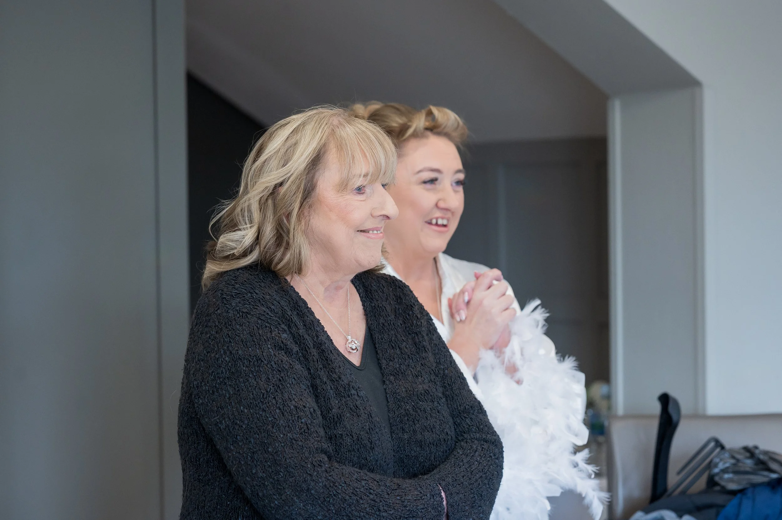 Two women standing side by side, both smiling and looking forward, in a modern indoor setting. The woman on the left has shoulder-length blonde hair, wears a black textured jacket, and a silver necklace. The woman on the right has short, light brown 