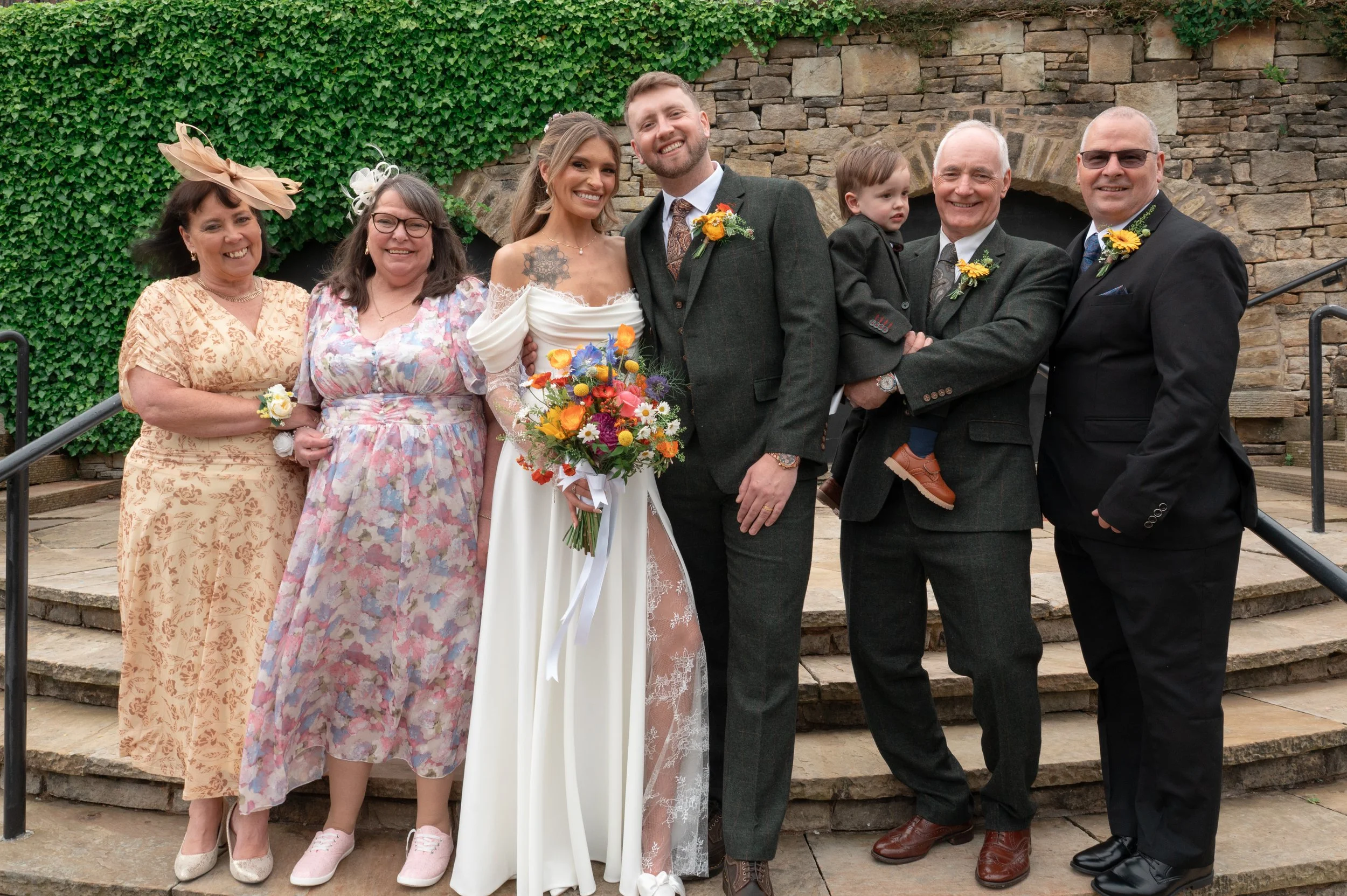 Group of seven people, including a bride and groom, standing on stone steps in front of a brick wall and greenery at a wedding celebration.