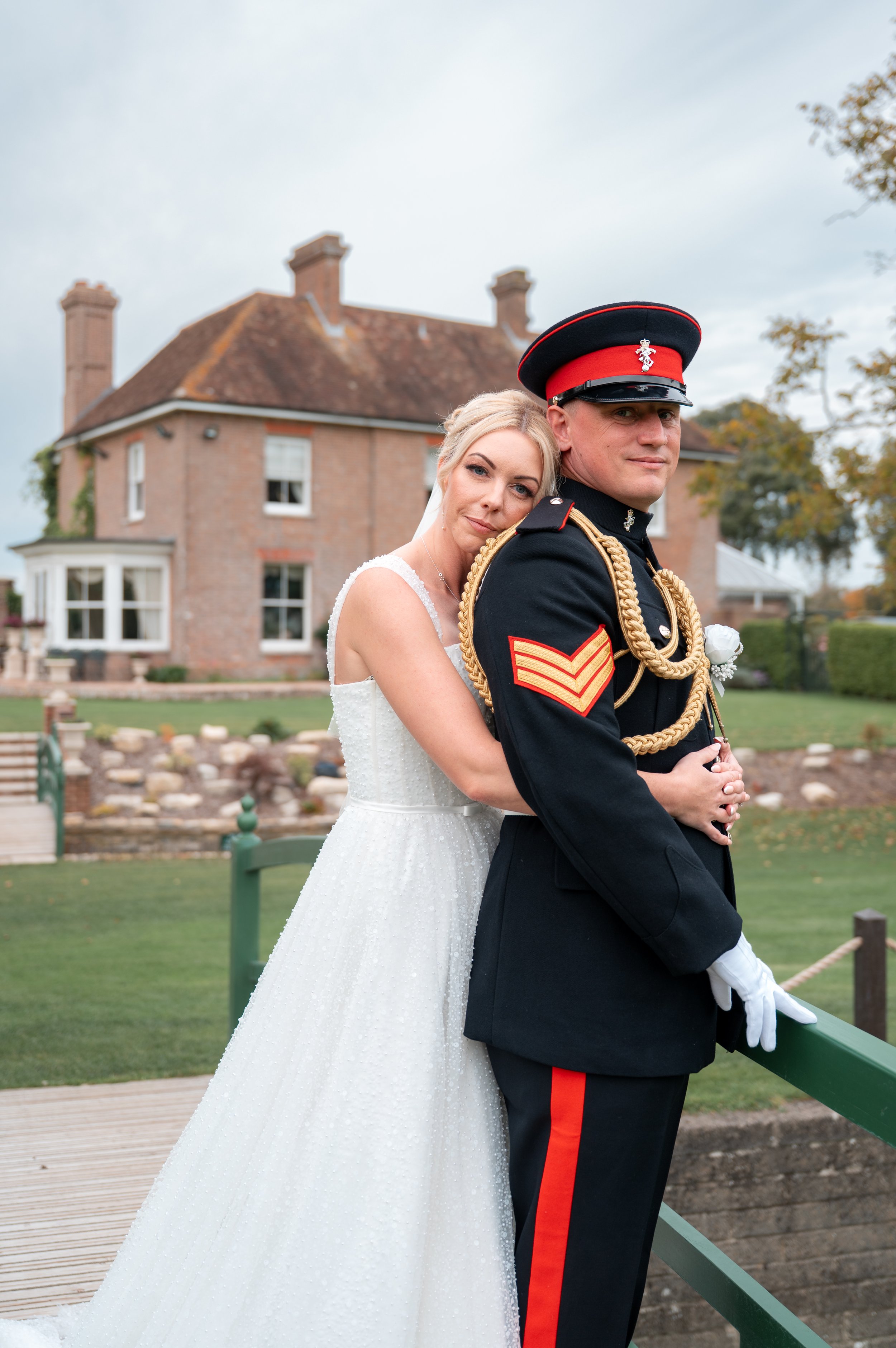 A bride and groom in wedding attire standing outdoors near a historic brick building and a pond. The bride is leaning against the groom, with her head resting on his shoulder. The groom is in a military uniform with a peaked cap with a red band and badge, and decorative gold cords on his uniform. The bride is wearing a white wedding dress and has blonde hair, while the groom has short dark hair. They are both smiling softly, creating a romantic scene.