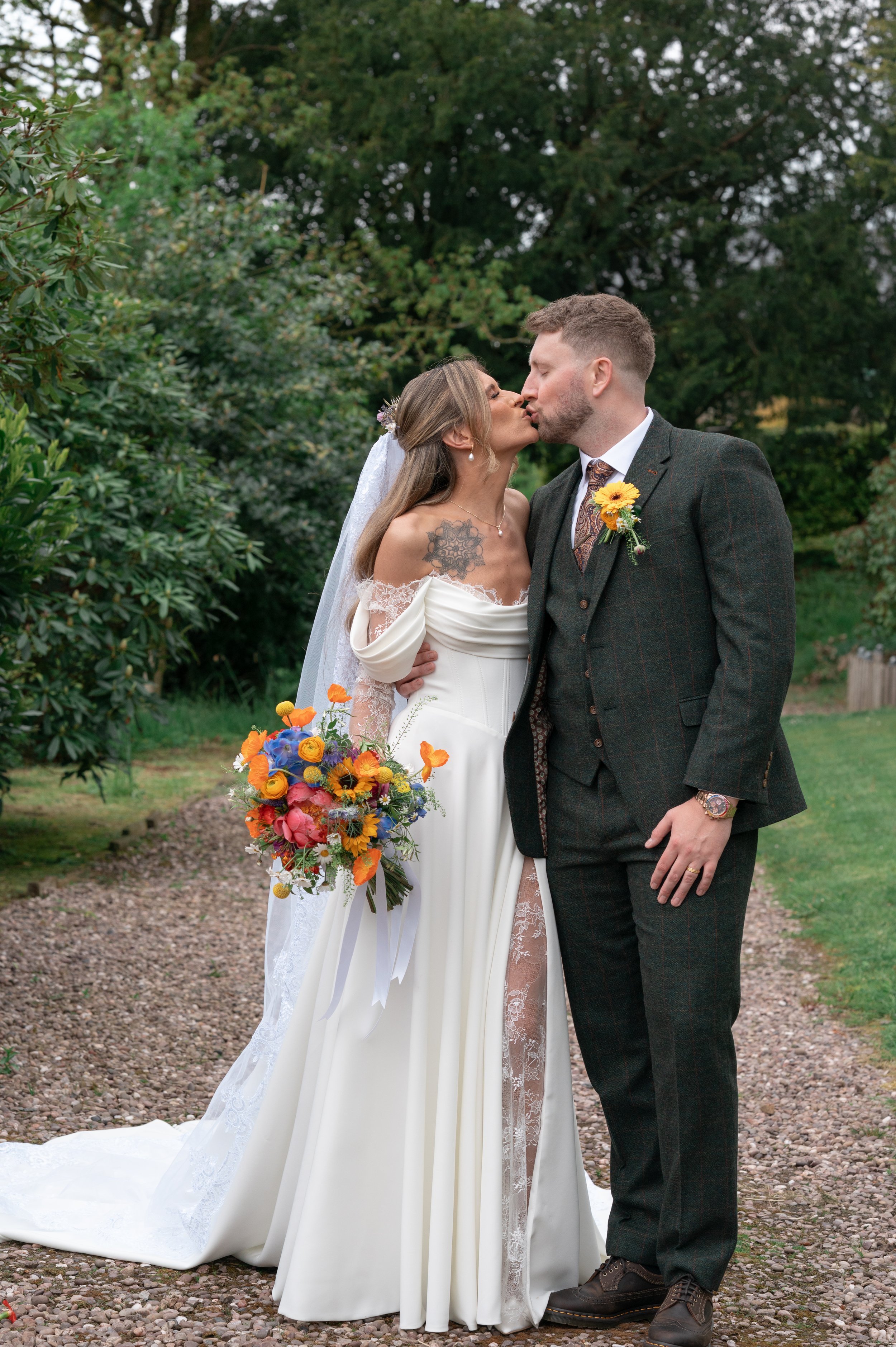 A bride and groom kissing outdoors during their wedding, with lush green trees in the background. The bride is wearing a white off-shoulder wedding dress with lace sleeves and a veil, holding a colorful bouquet. The groom is dressed in a dark plaid s