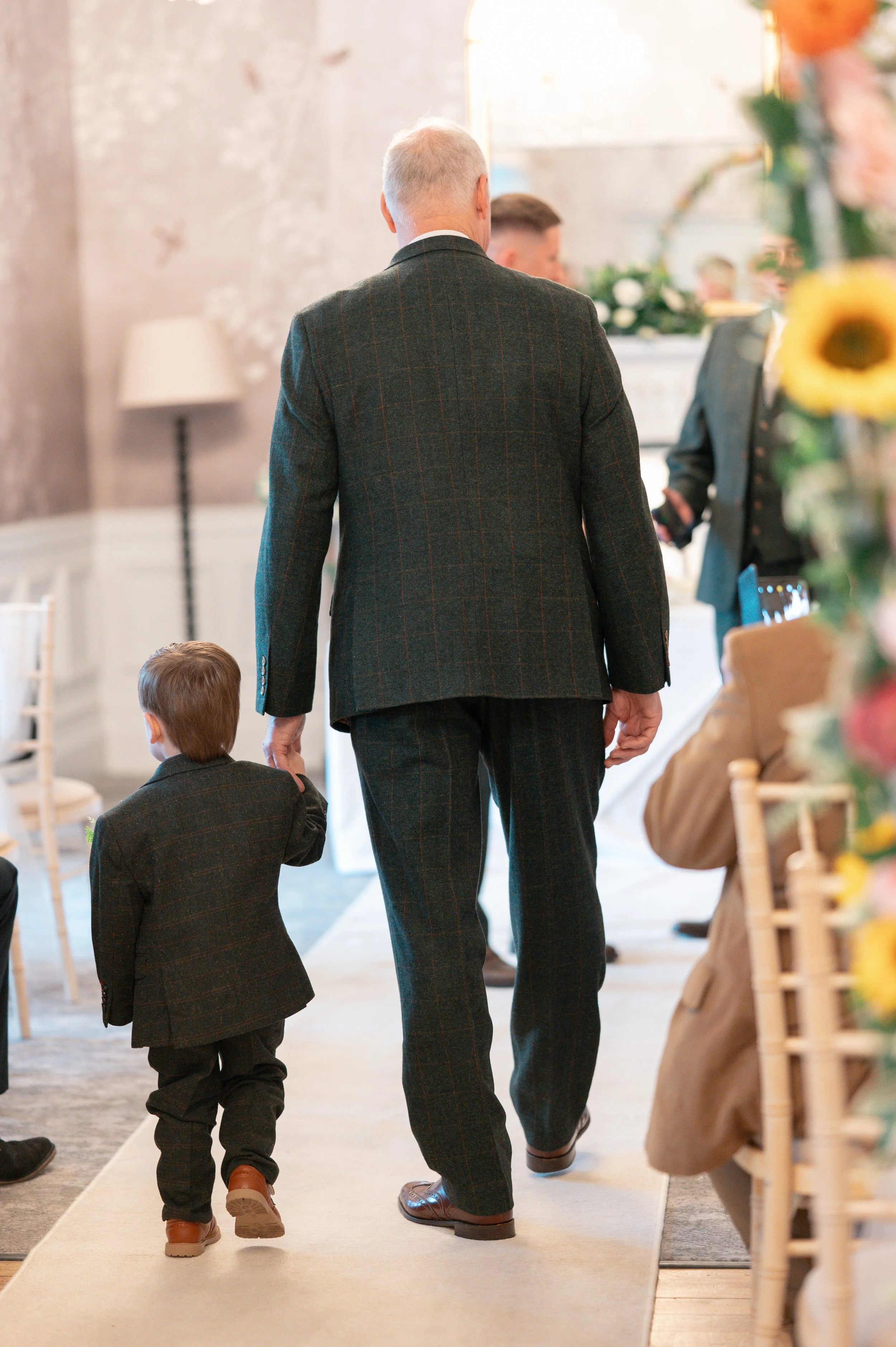 Man and young boy walking down an aisle at a formal indoor event, with the man holding the boy's hand. The man is wearing a dark checked suit, and the boy is also dressed in a dark suit. The background shows other people and floral decorations.