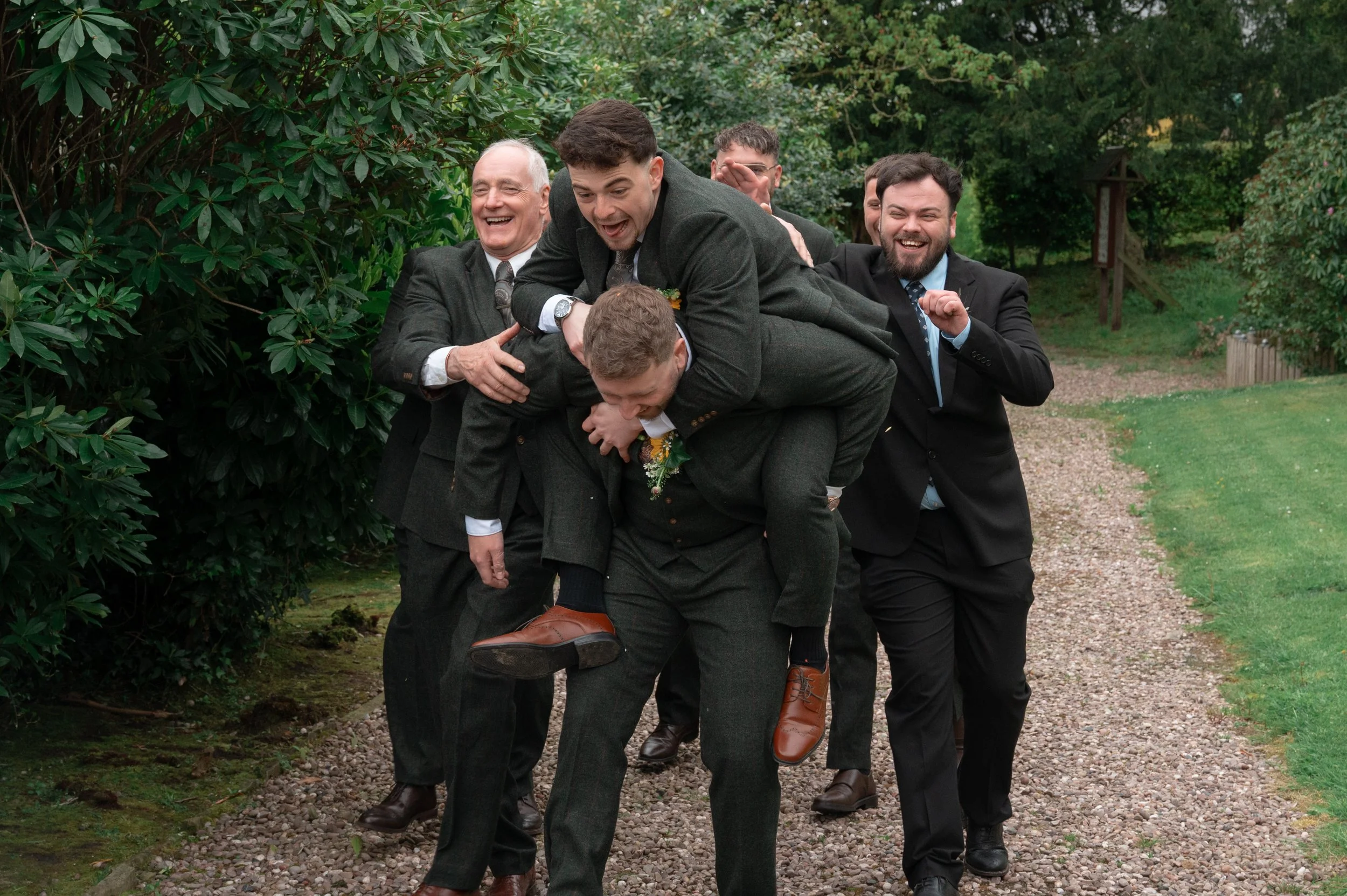 Group of men in suits arranged in a human train, outdoors during daytime, surrounded by greenery.