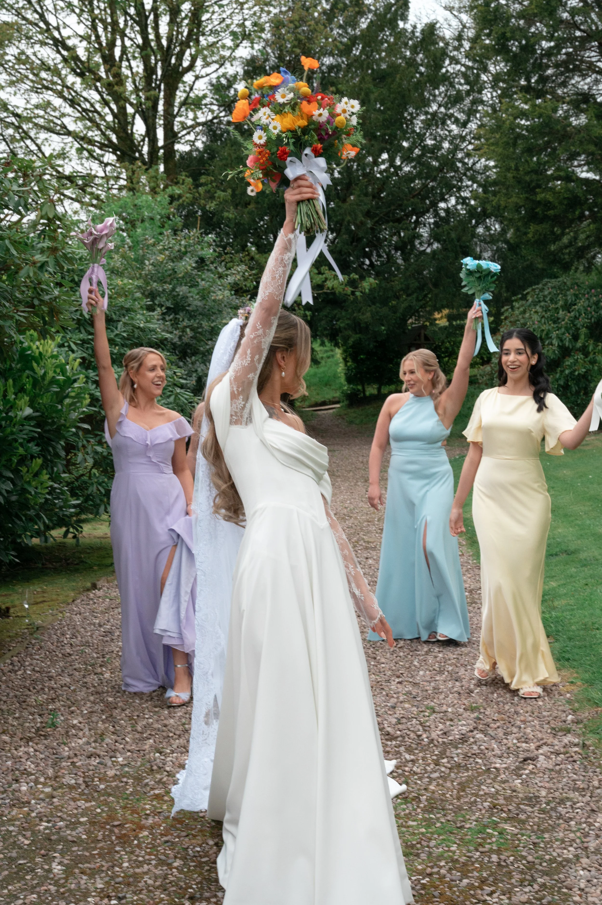 A bride in a white wedding dress holds a bouquet of orange, yellow, white, and blue flowers high, while four bridesmaids in pastel-colored dresses follow behind her, all celebrating outdoors on a gravel pathway surrounded by greenery and trees.