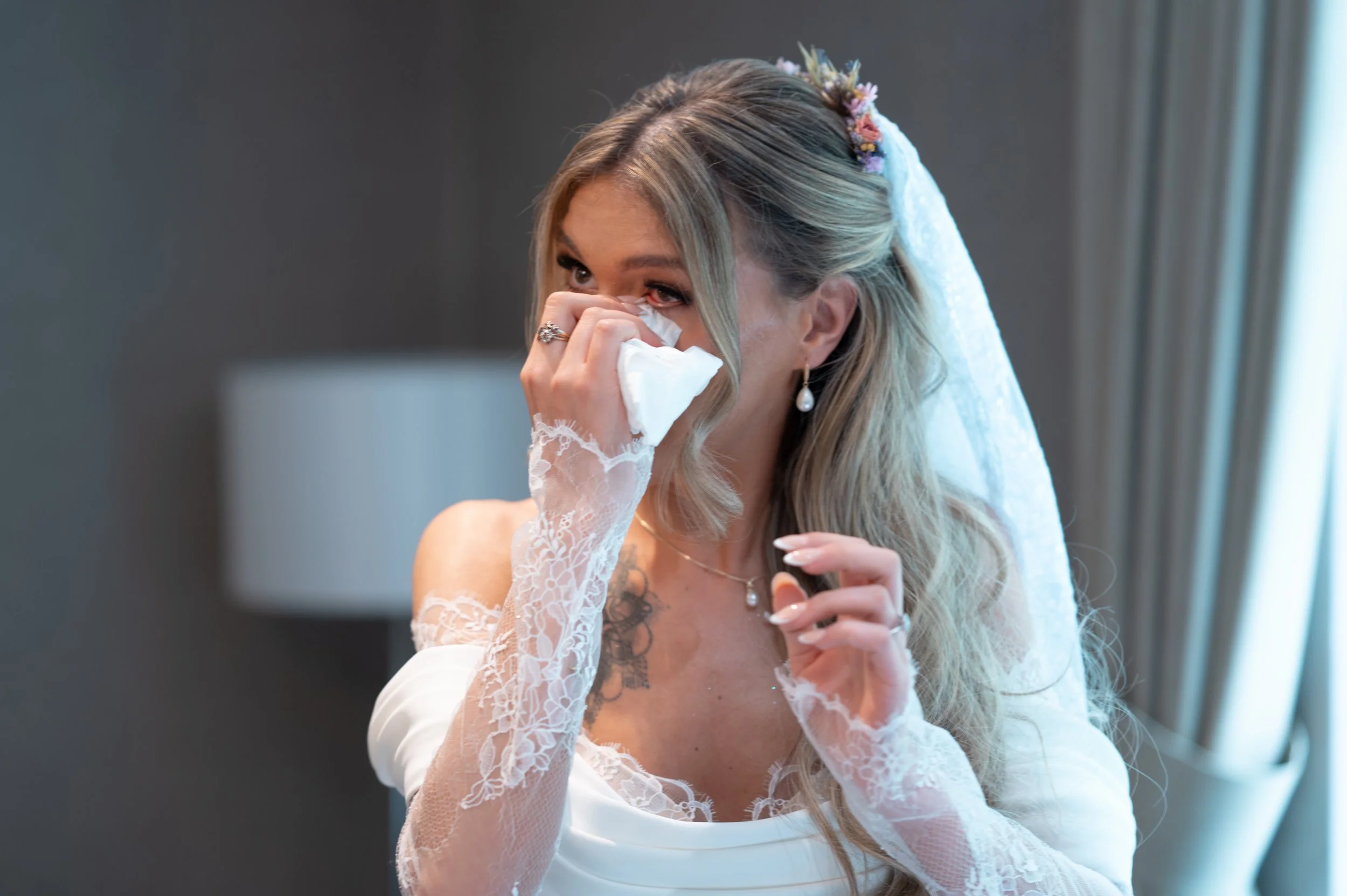 A bride with blonde hair and a lace wedding dress is crying and wiping her eyes with tissue. She is wearing pearl earrings, a ring, and a necklace, and has flowers in her hair.