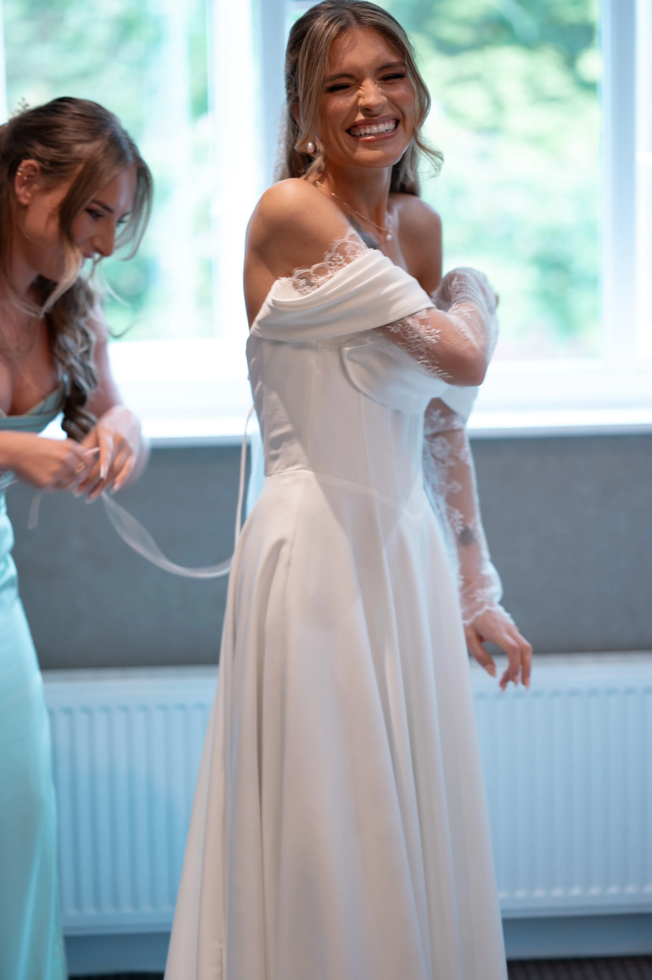 A woman in a white wedding dress smiling and adjusting her dress, with another woman helping her, in front of a large window with greenery outside.