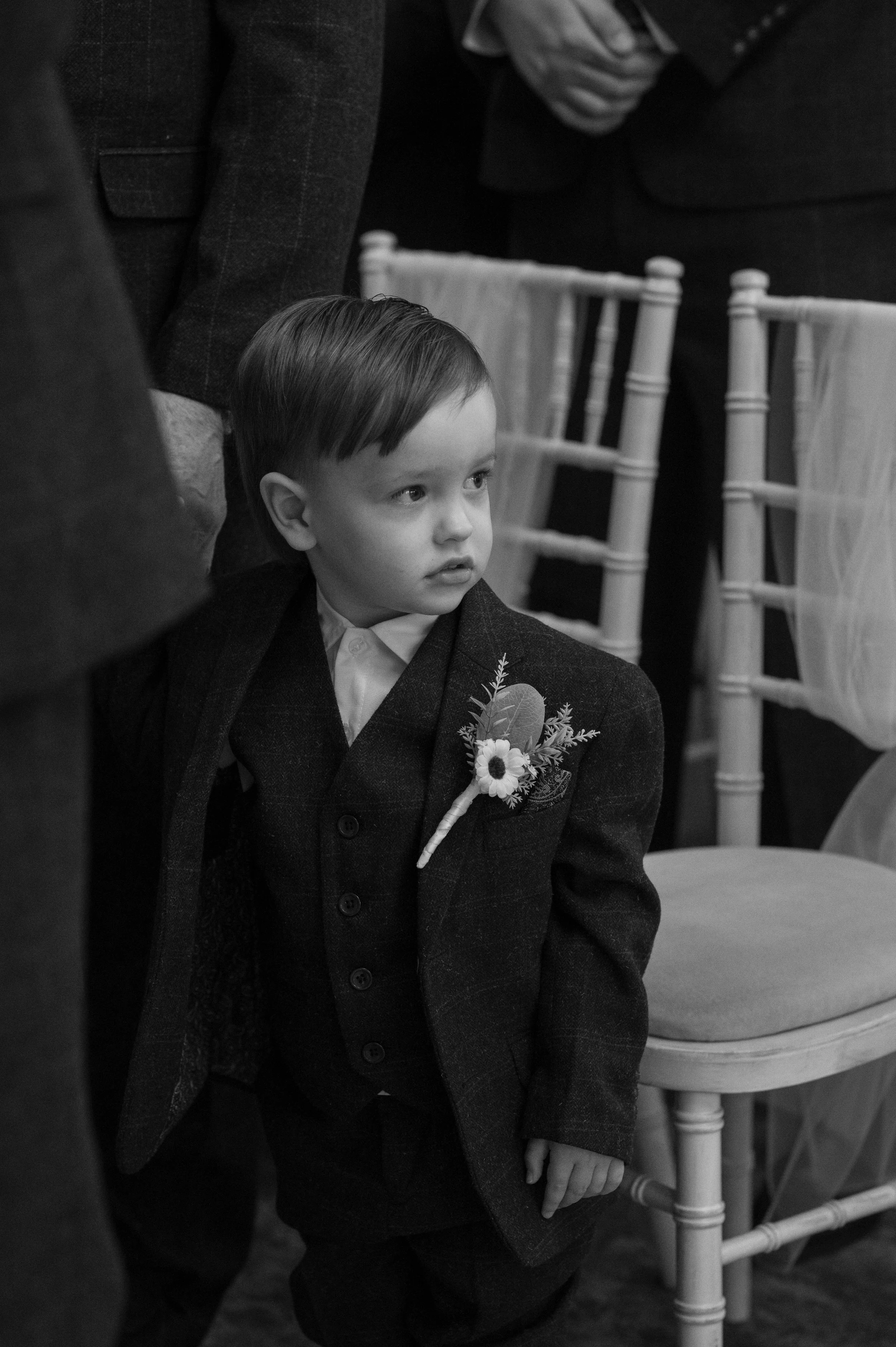 A young boy in formal attire with a boutonnière on his suit jacket, standing in a room with chairs, possibly during a wedding or formal event.