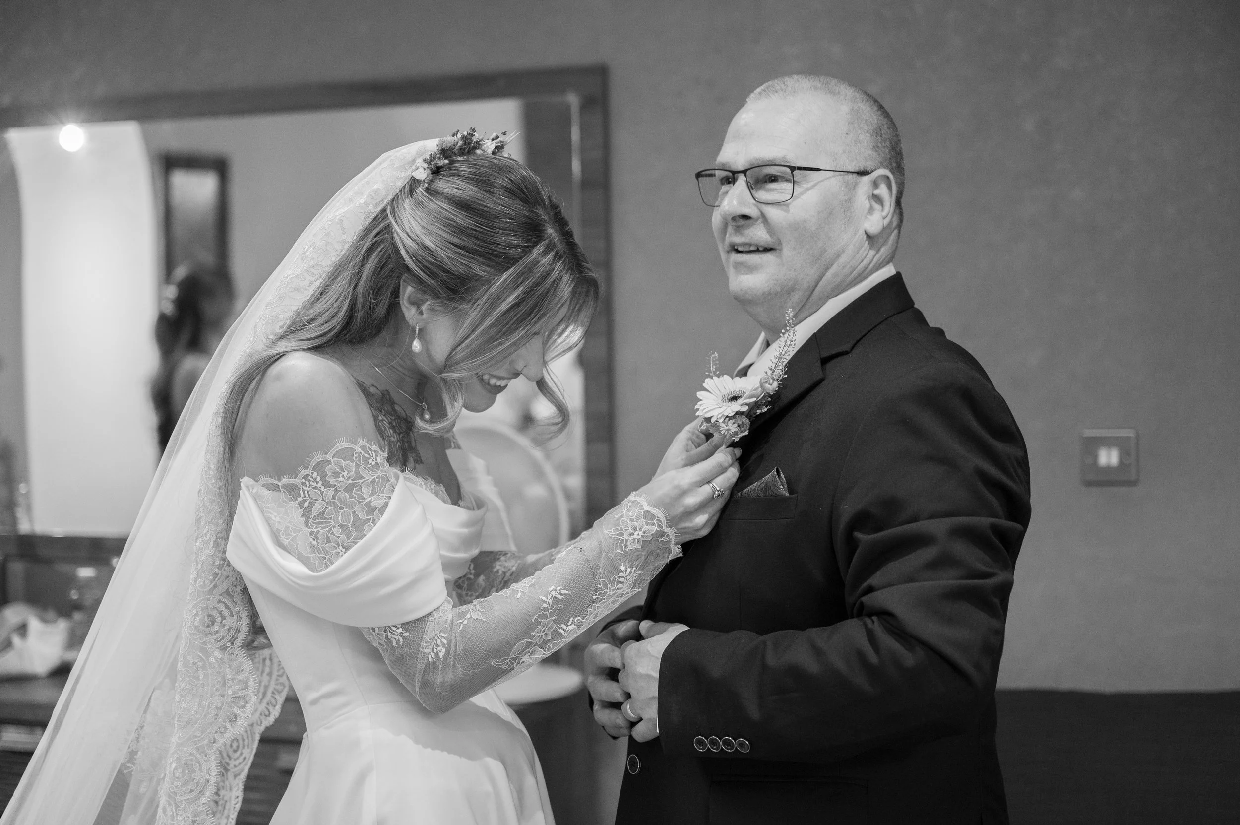 A bride in a lace wedding gown and veil smiles and leans her head down towards a groom who is dressed in a suit with a boutonniere, smiling and looking at her during a wedding ceremony.