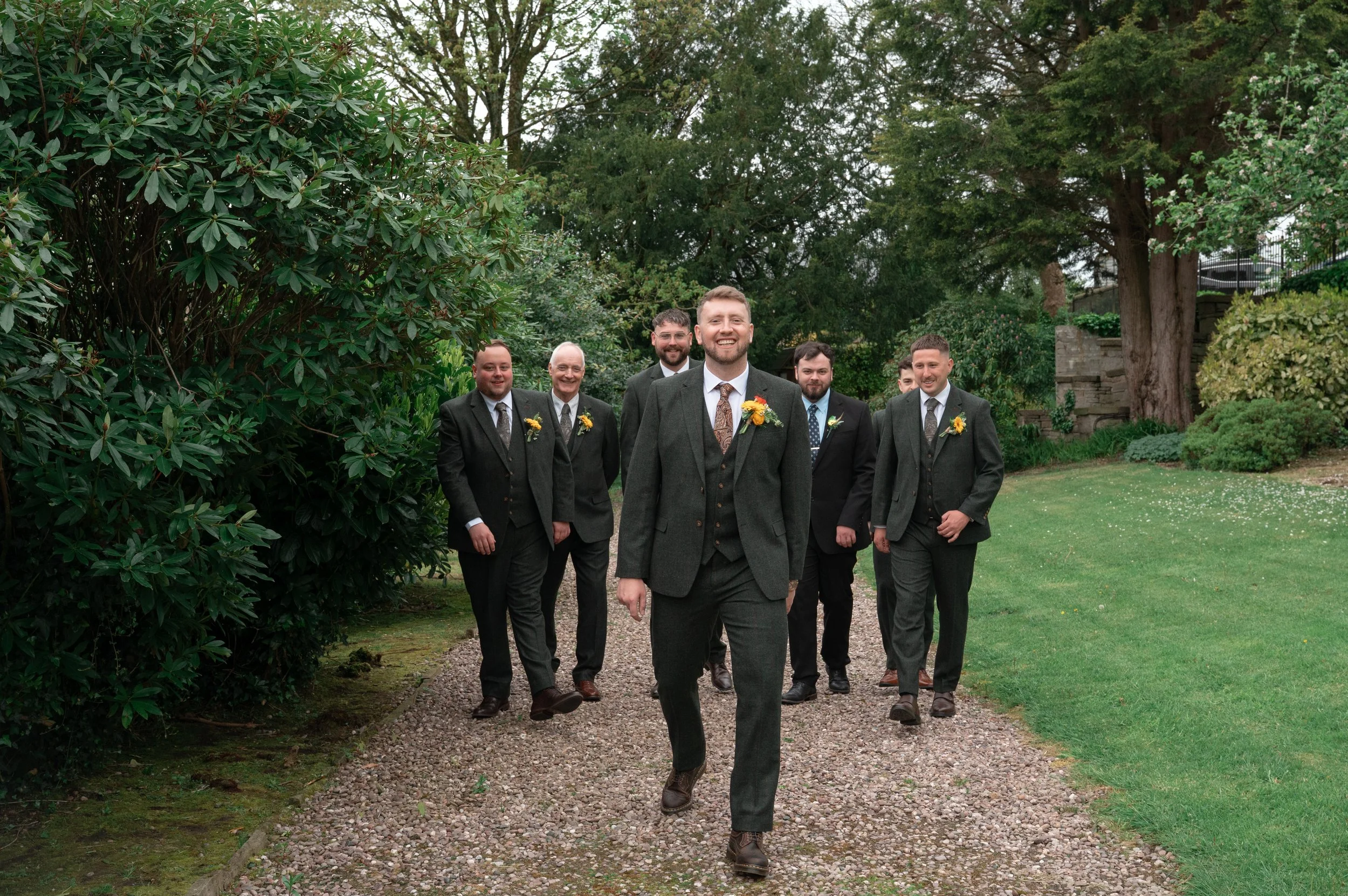 A group of seven men in suits walking along a gravel path in a garden with lush trees and bushes, smiling and looking happy.