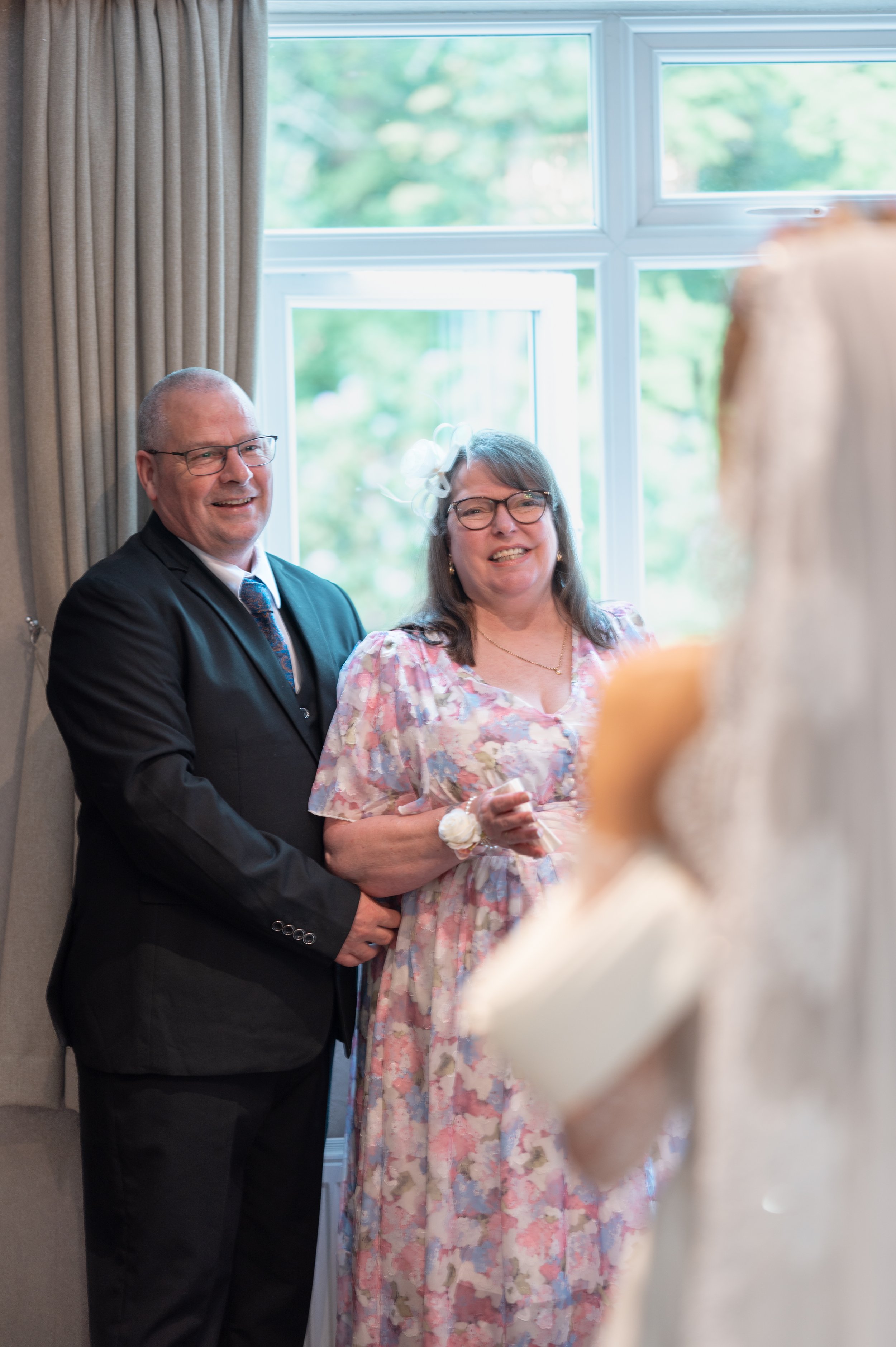 Smiling couple dressed formally, with the man in a black suit and the woman in a floral dress and glasses, standing by a window in a well-lit room during a social event.