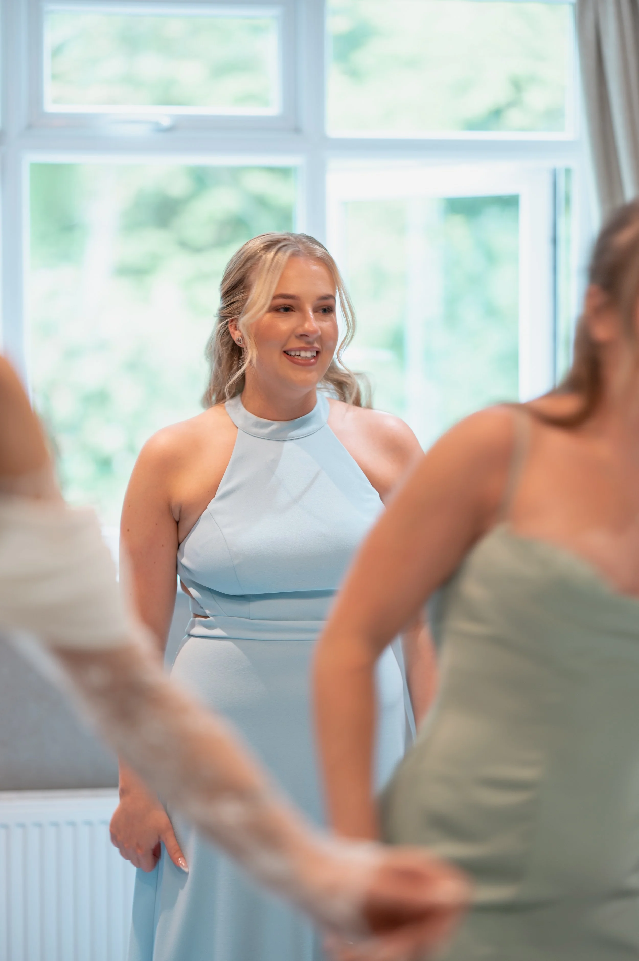 A group of women dressed in pale blue dresses participating in a wedding ceremony, with a woman in focus smiling and talking, standing in front of a large window with a view of green trees outside.