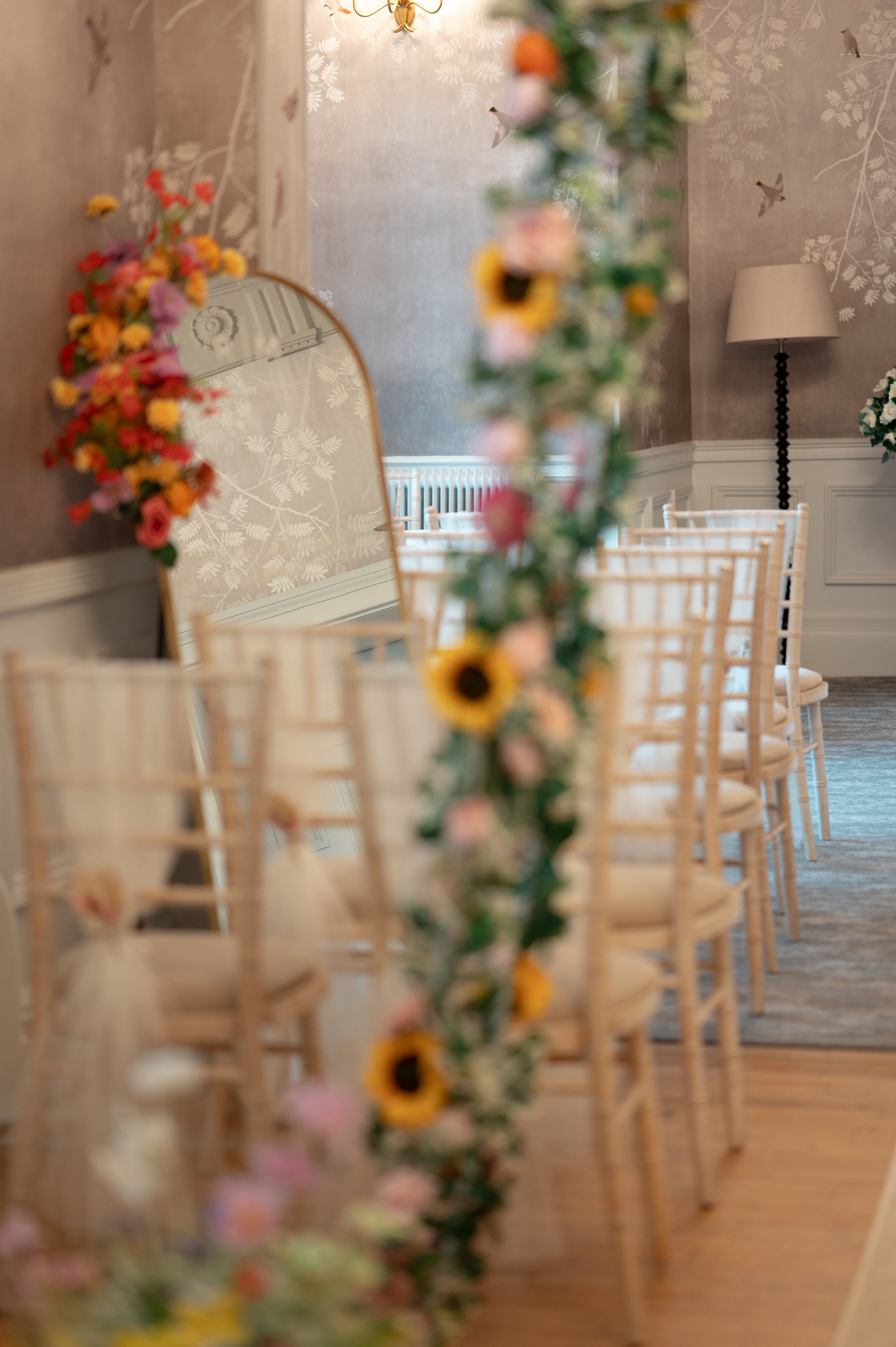 A blurry image of a room with a floral curtain, a large mirror decorated with flowers, a chandelier, a wall with a bird pattern, a tall floor lamp, and chairs with white cushions.