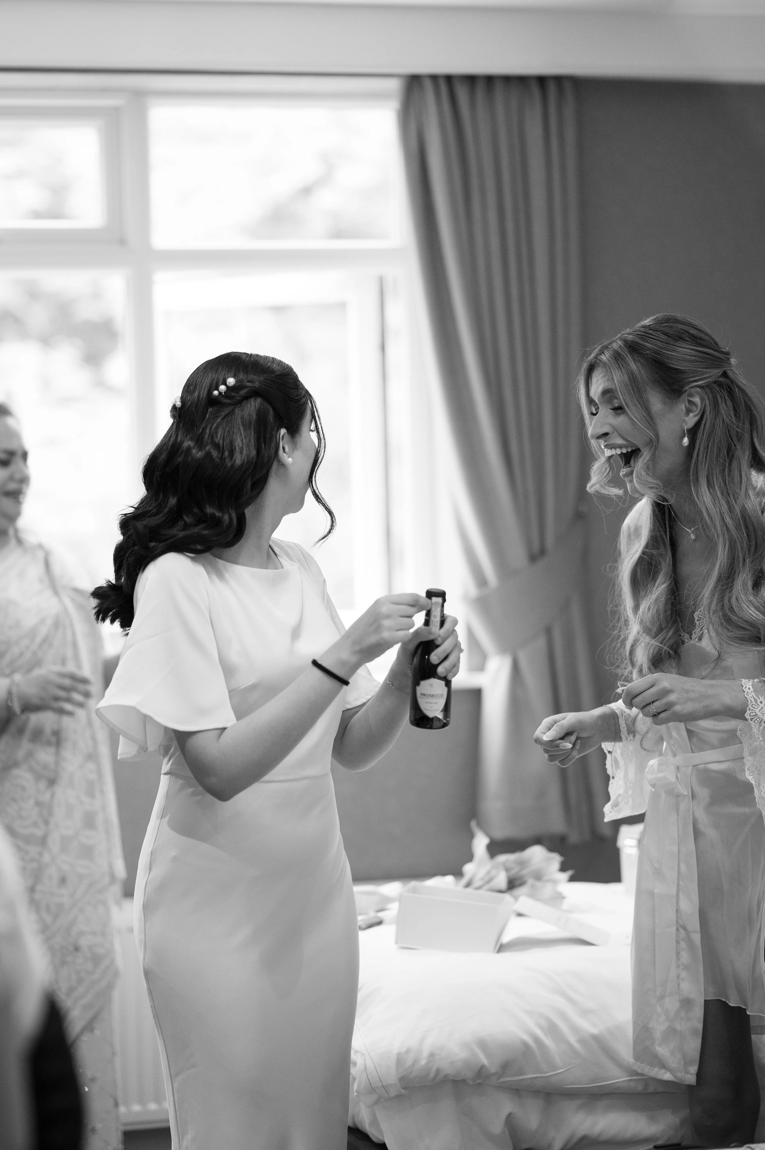Women in wedding dresses laughing and talking indoors by a bed, with sunlight coming through large windows and curtains.