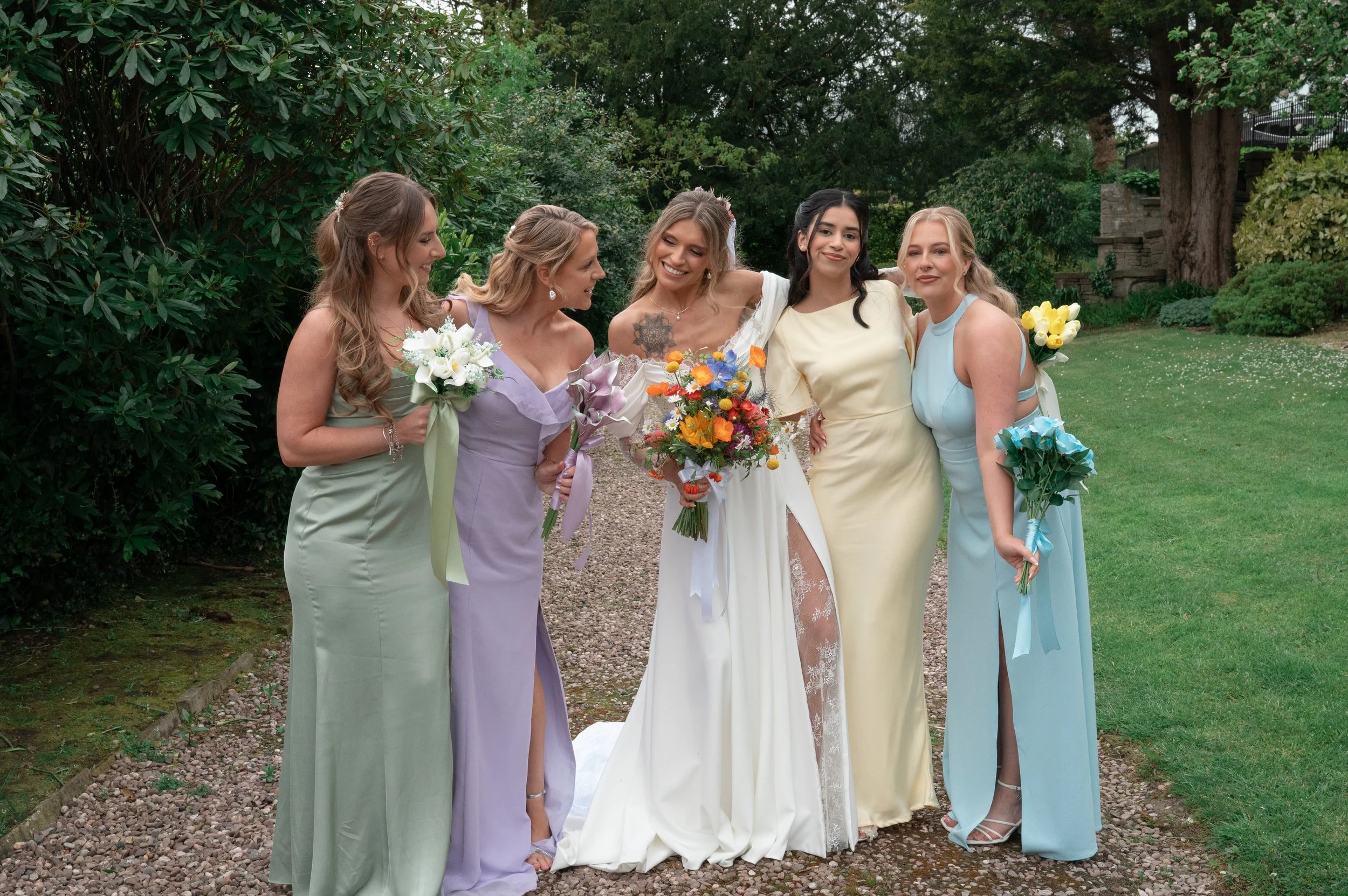 A bride and five women dressed in pastel-colored gowns standing on a gravel path outdoors surrounded by greenery, holding bouquets of flowers, at a wedding.