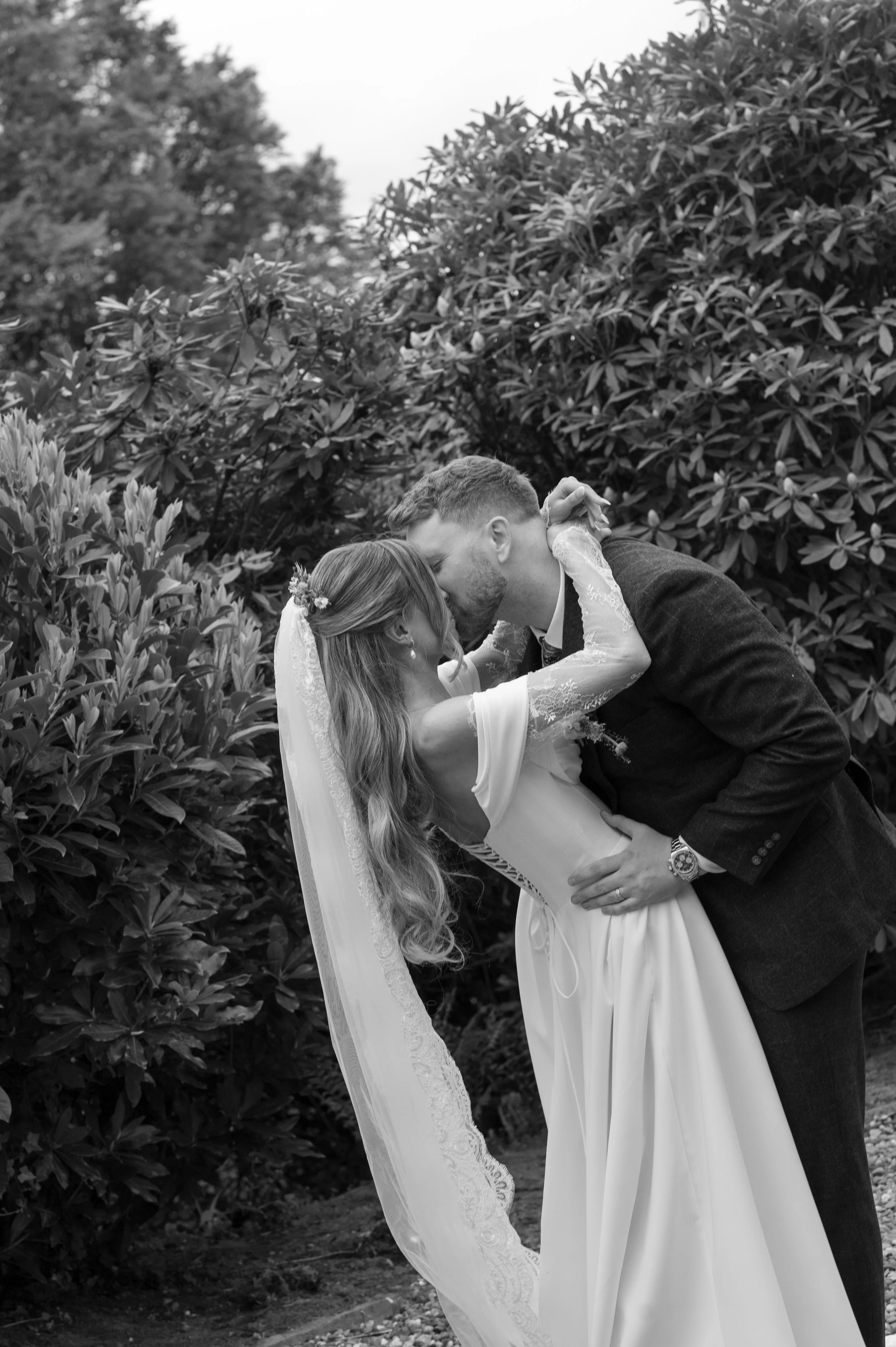 A newlywed couple sharing a kiss outdoors, surrounded by bushes and trees, in black and white.