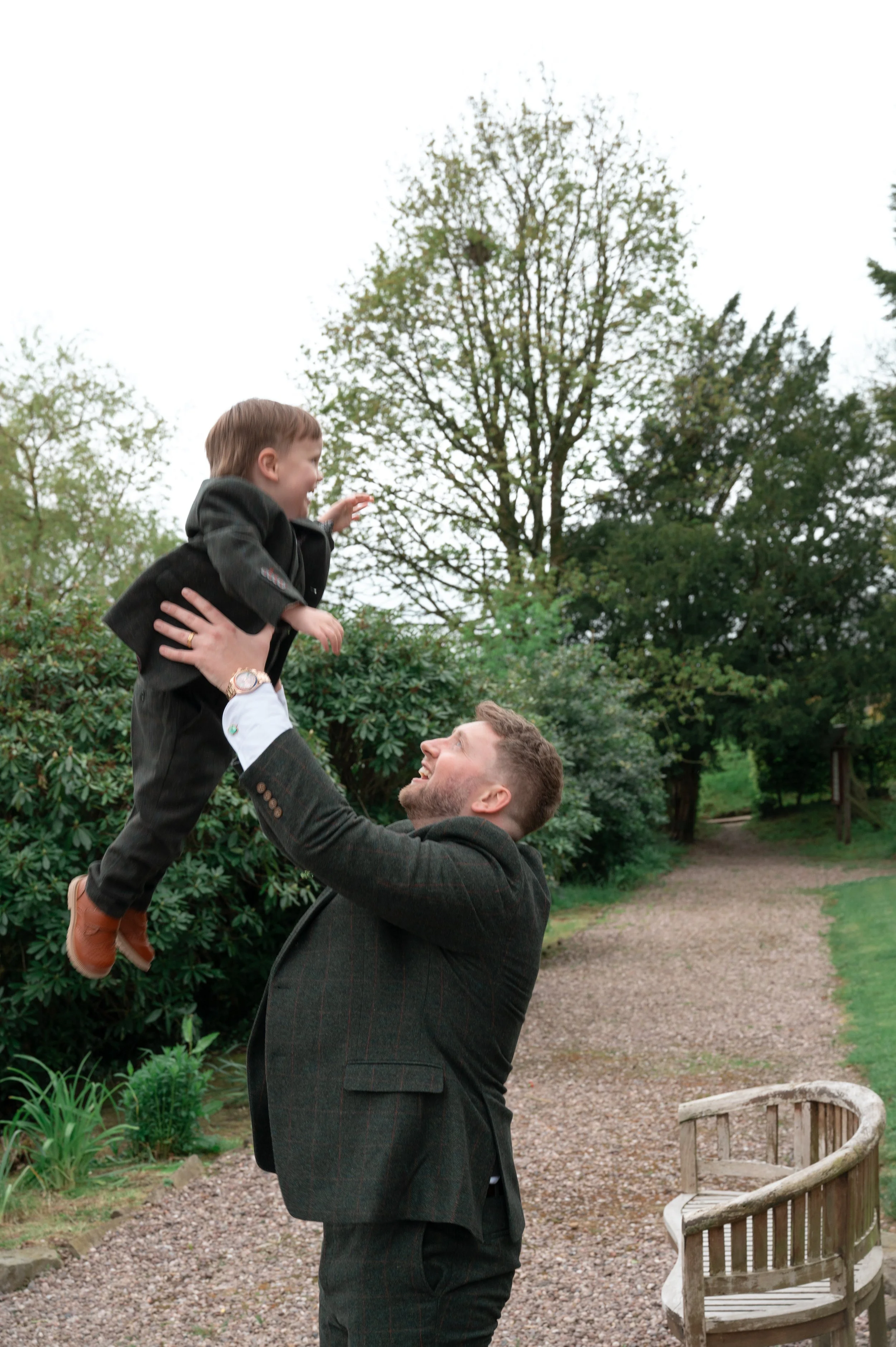 A man wearing a dark suit is lifting a young boy wearing a black coat, white shirt, and brown shoes in a park. The boy is smiling and reaching out with his right hand. There are trees and greenery in the background.