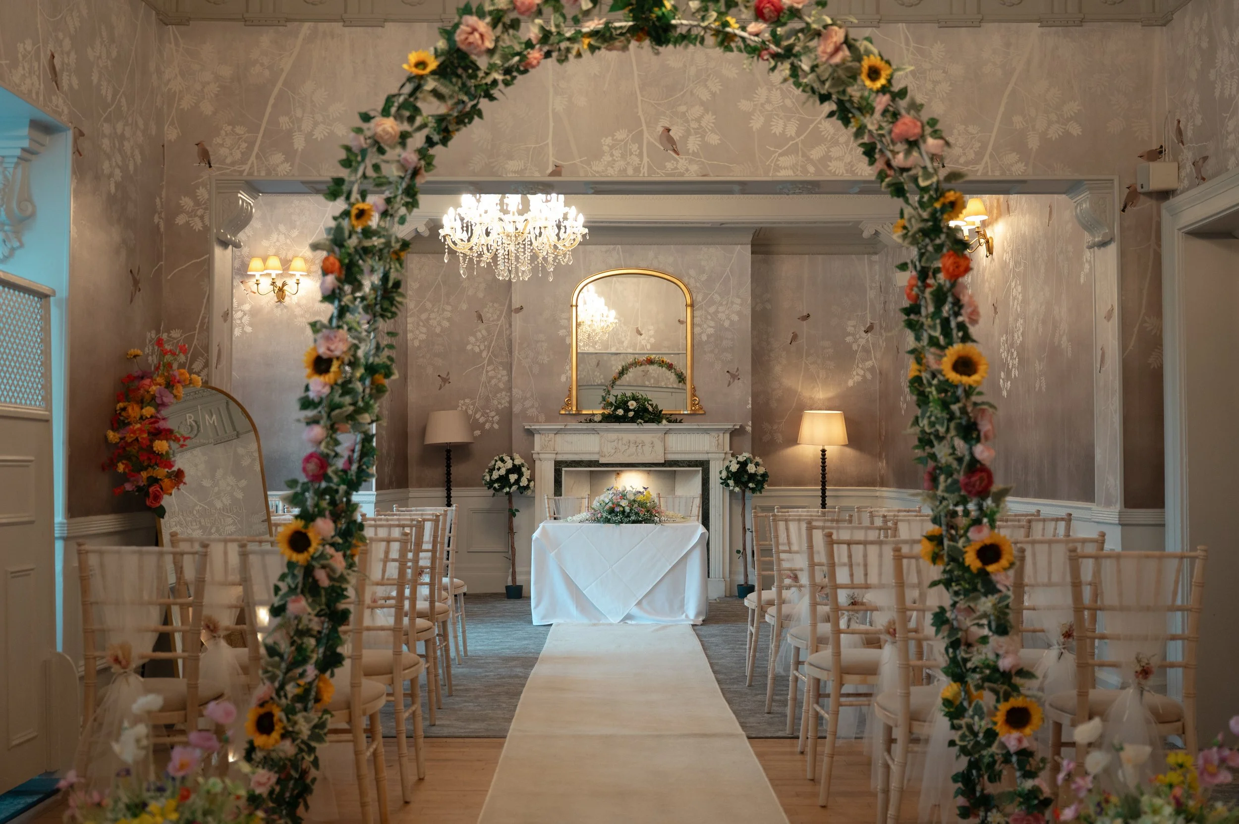 Indoor wedding setup with a carpeted aisle, floral arch, chairs, and a decorated table at the front with a mirror and chandelier overhead.