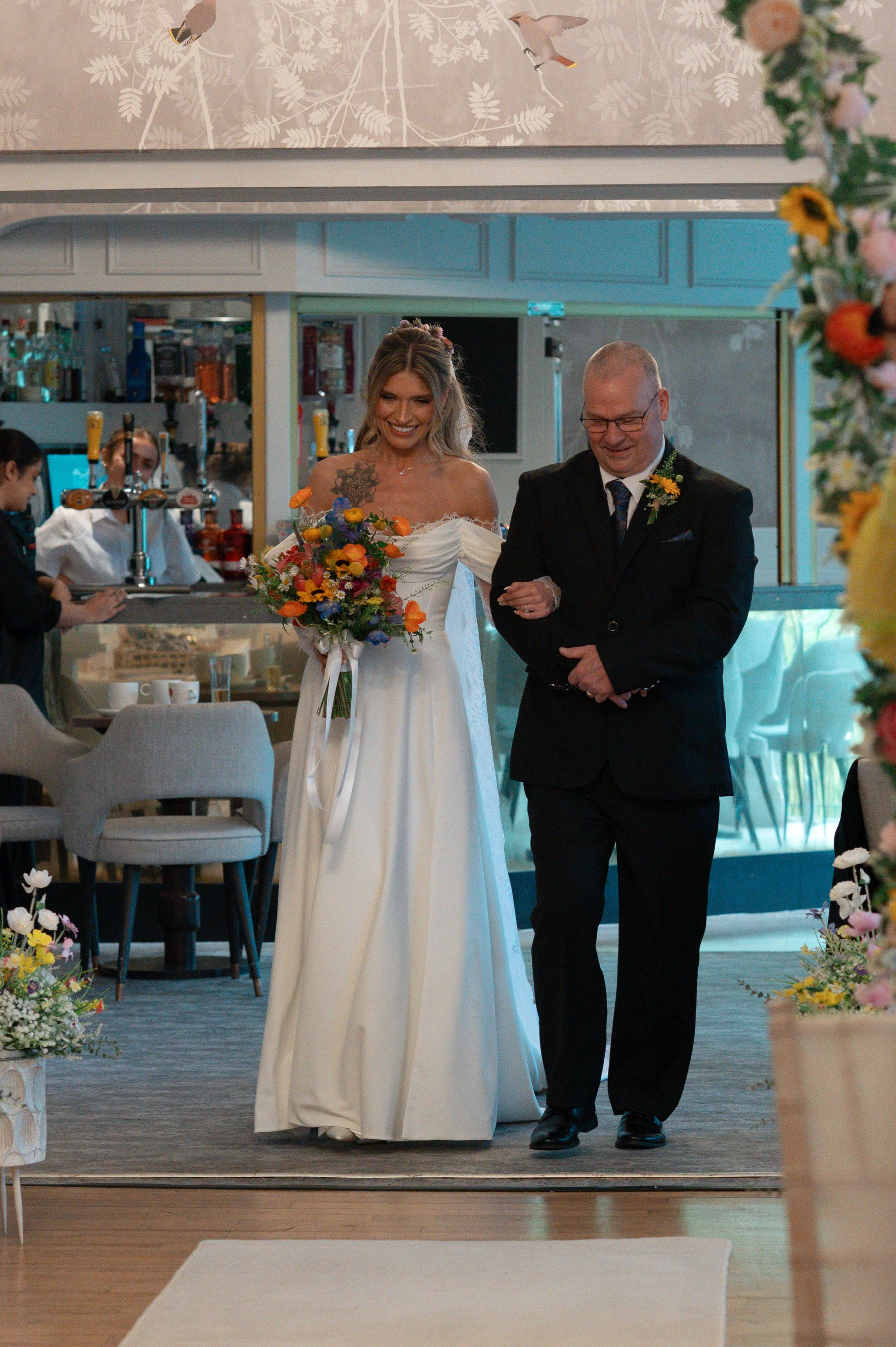 A bride in a white wedding gown holding a colorful bouquet, and a man in a black suit with a boutonniere, walking together inside a decorated venue.