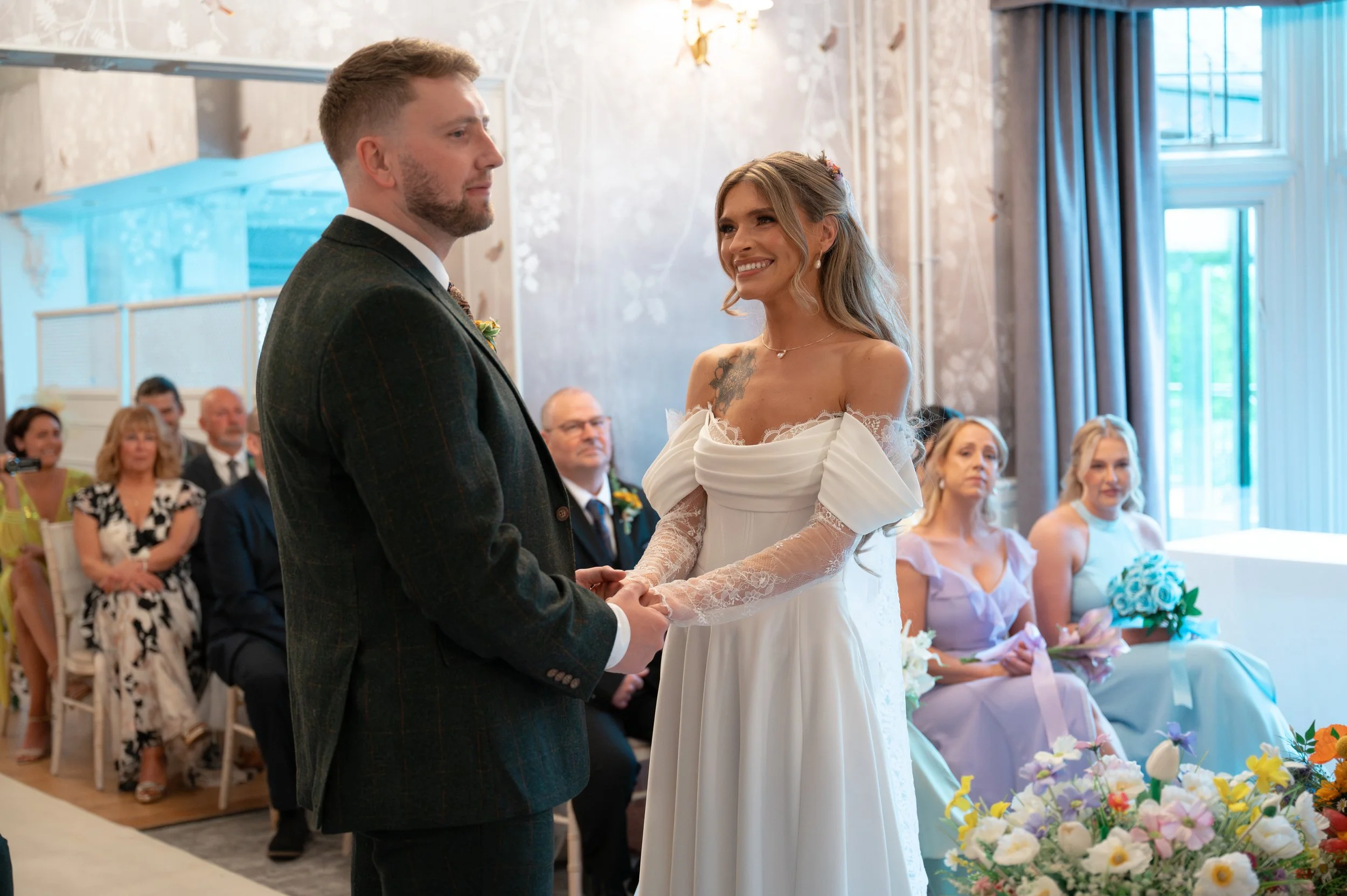 A bride and groom holding hands during their wedding ceremony, with guests seated behind them in a decorated indoor venue.