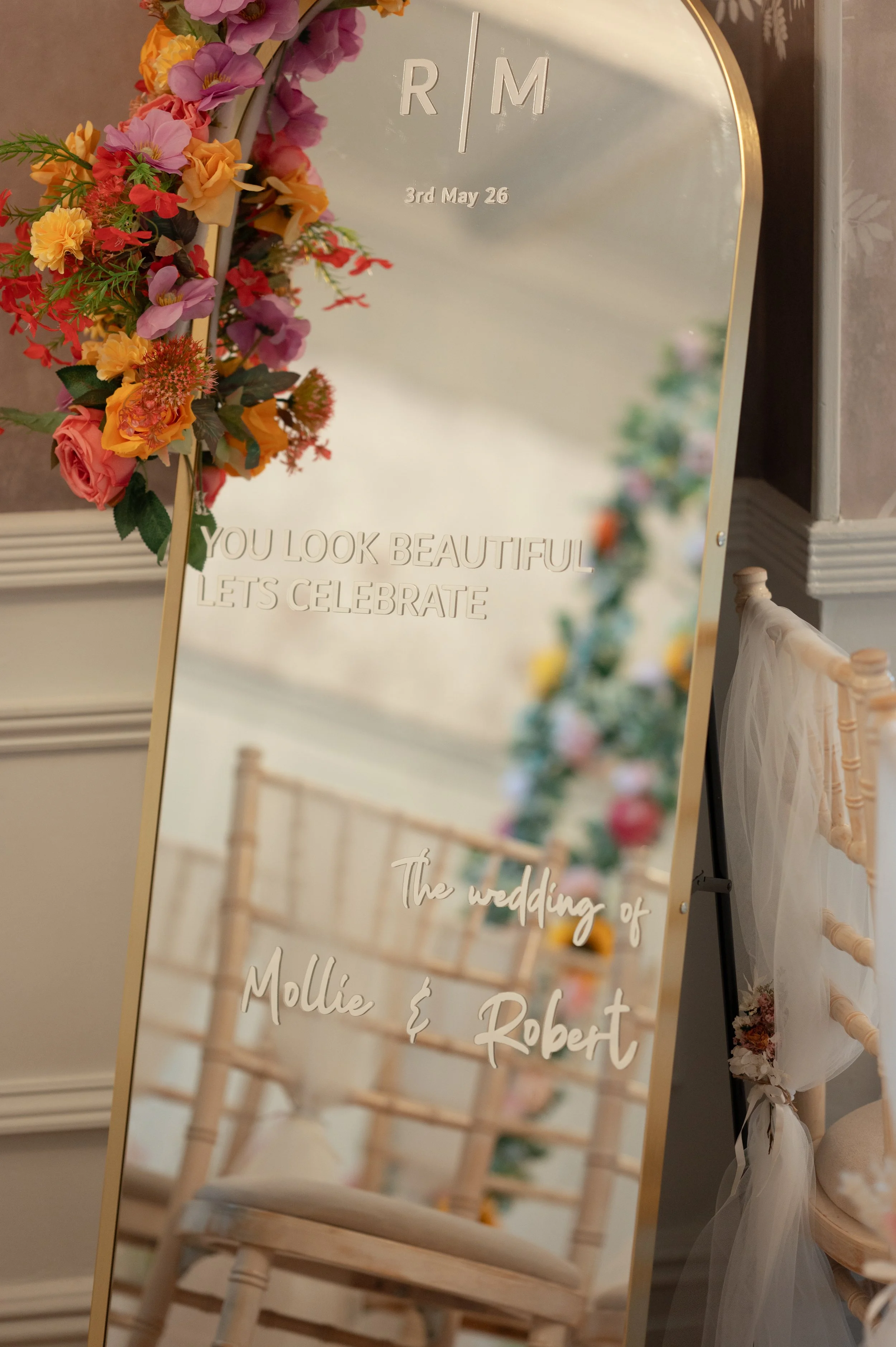 A decorated wedding mirror with floral arrangements on the upper left side, reflecting a white wooden chair with pink and white flowers in the background. The mirror has initials, date, and celebratory message engraved on it.