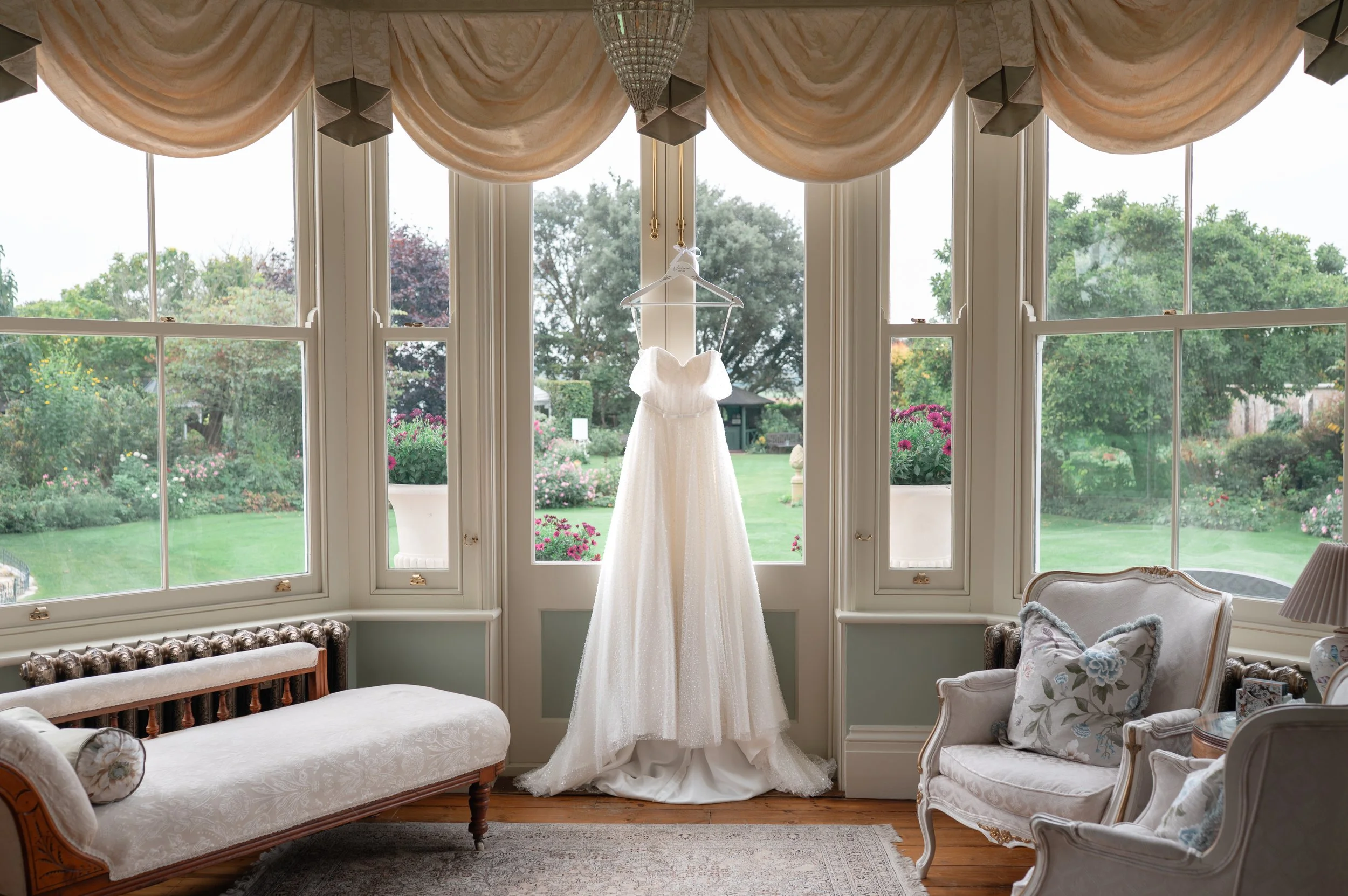 A white wedding dress hanging from a hanger in front of a bay window with curtains, overlooking a garden with trees and flowers.