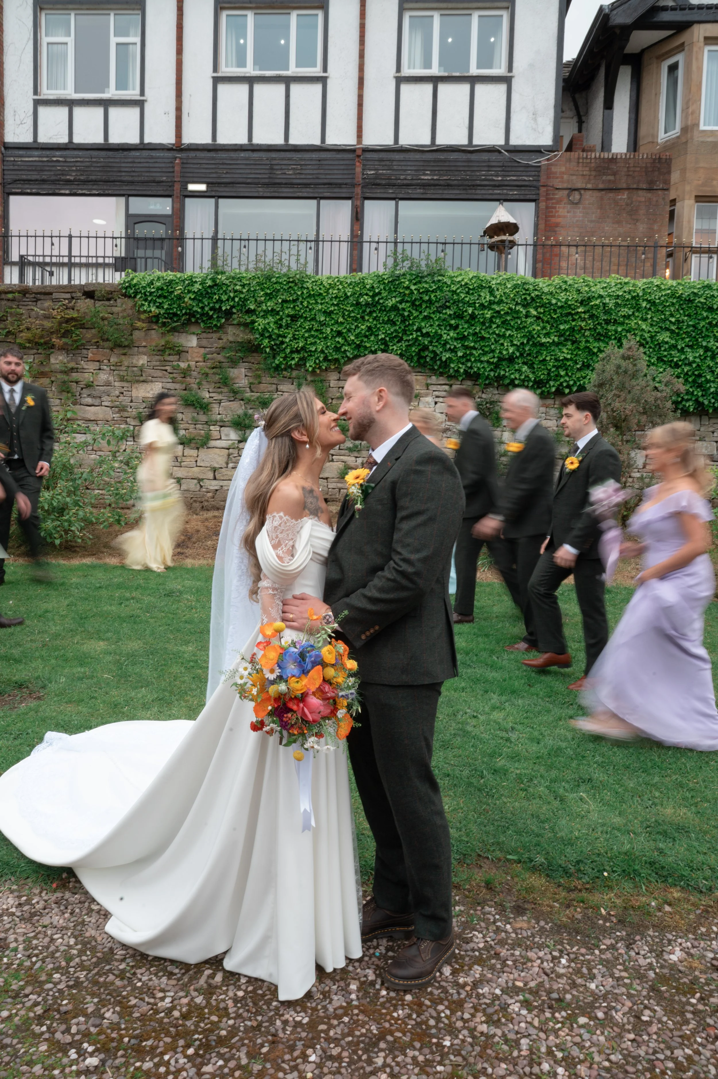 A bride and groom sharing a kiss during their wedding ceremony outdoors on a lawn, with wedding party members and guests in the background.