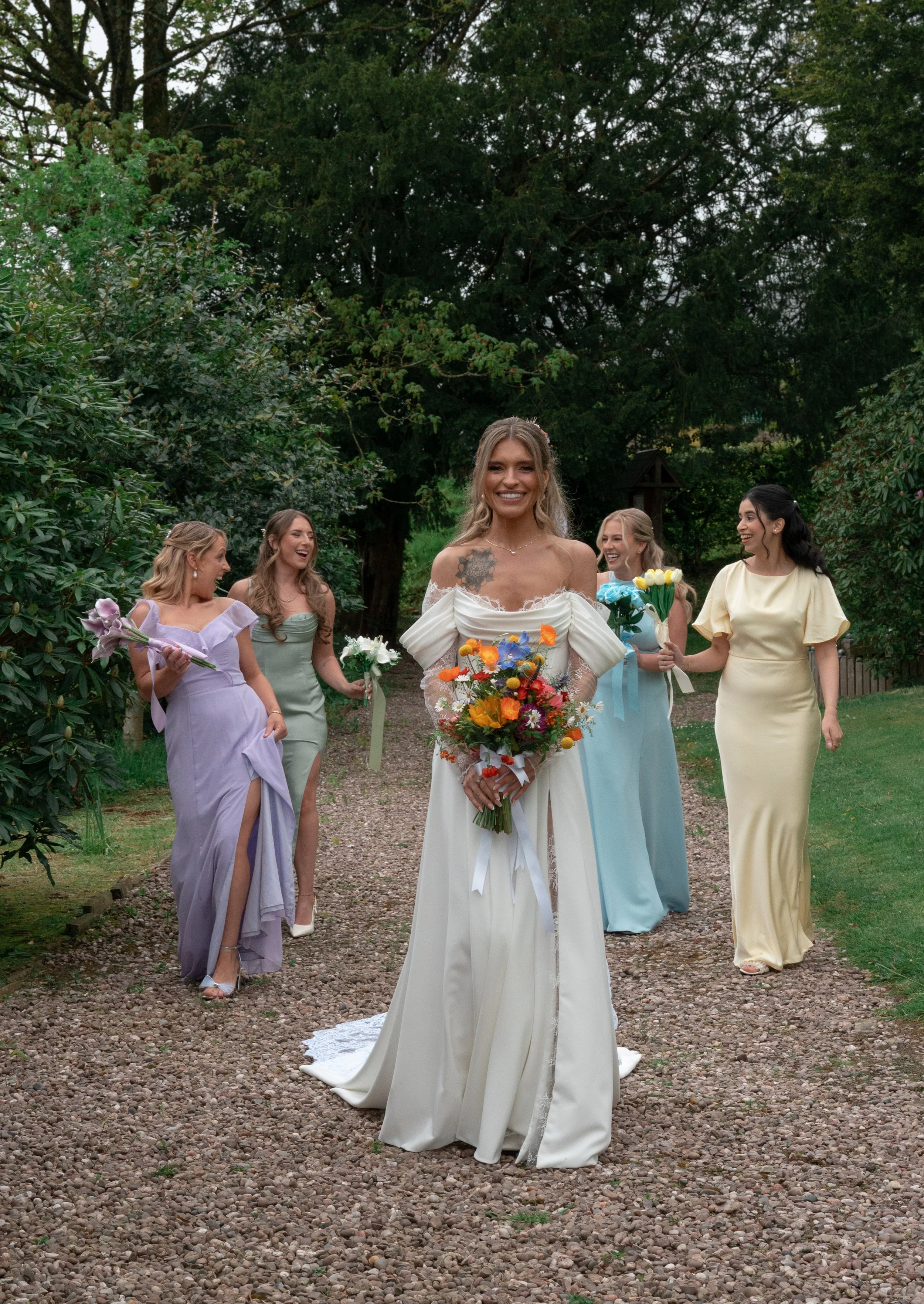 Bride in a white wedding dress holding a colorful bouquet, smiling, walking on a gravel path outdoors with five bridesmaids in pastel-colored dresses holding flowers, surrounded by green trees.