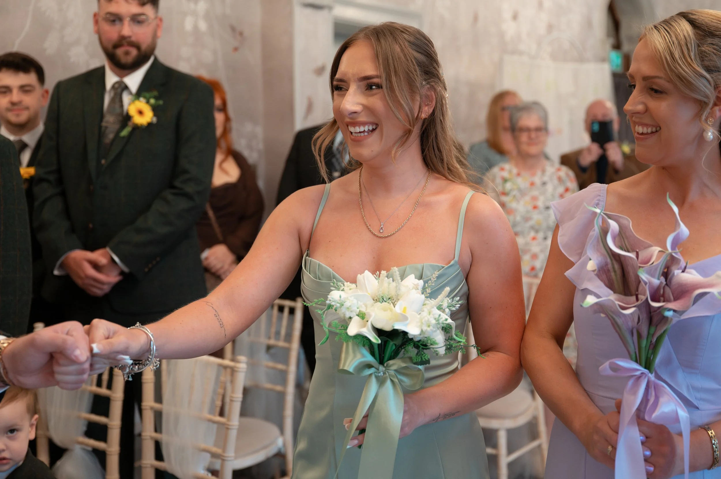 A smiling young woman in a light green dress holding a bouquet of white flowers, participating in a wedding ceremony, with other guests and an ornate wall in the background.