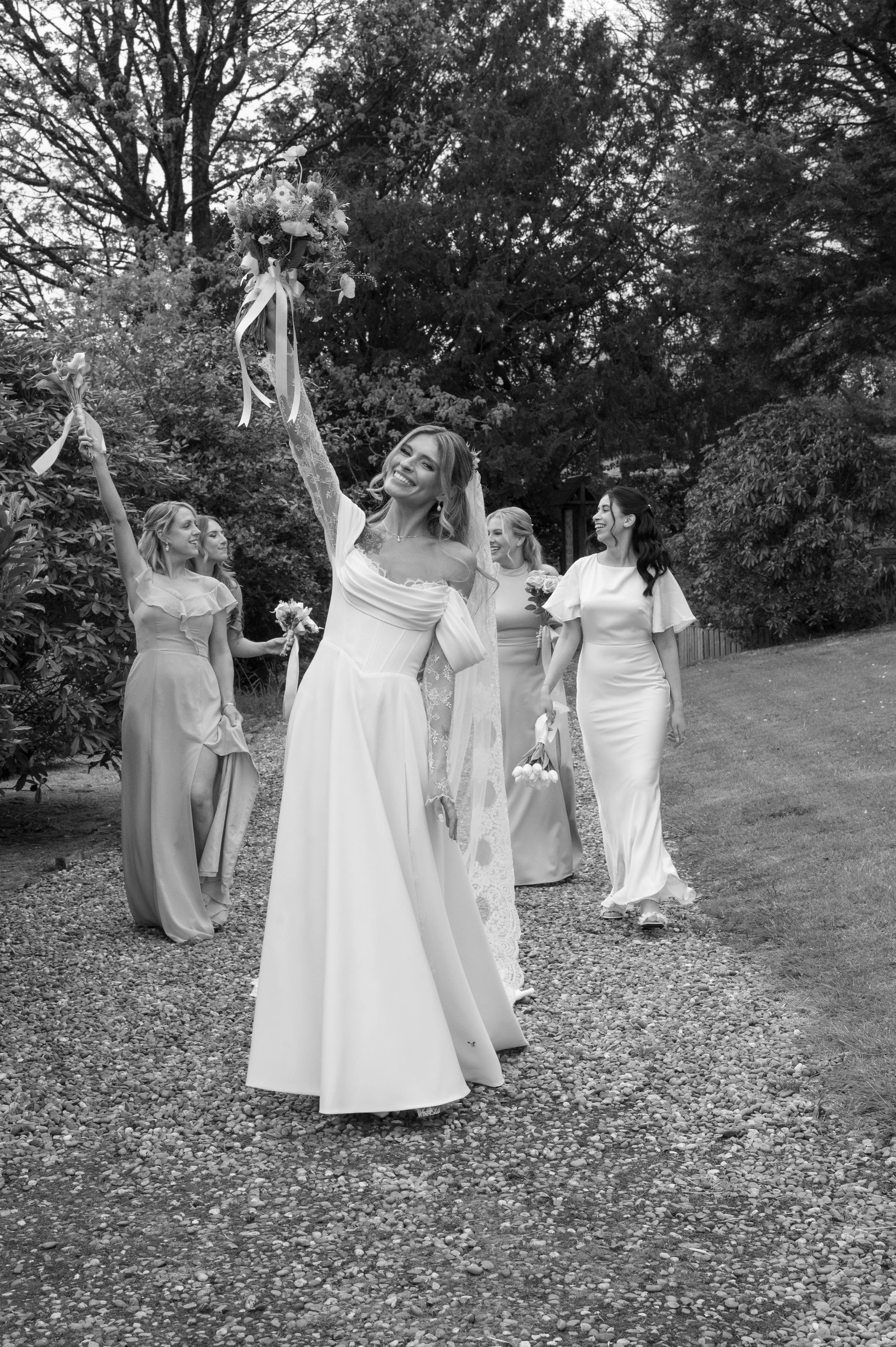 A woman in a wedding dress holds a bouquet of flowers with ribbons high above her head, smiling. Four women in matching dresses walk behind her, holding bouquets, in an outdoor setting surrounded by trees.