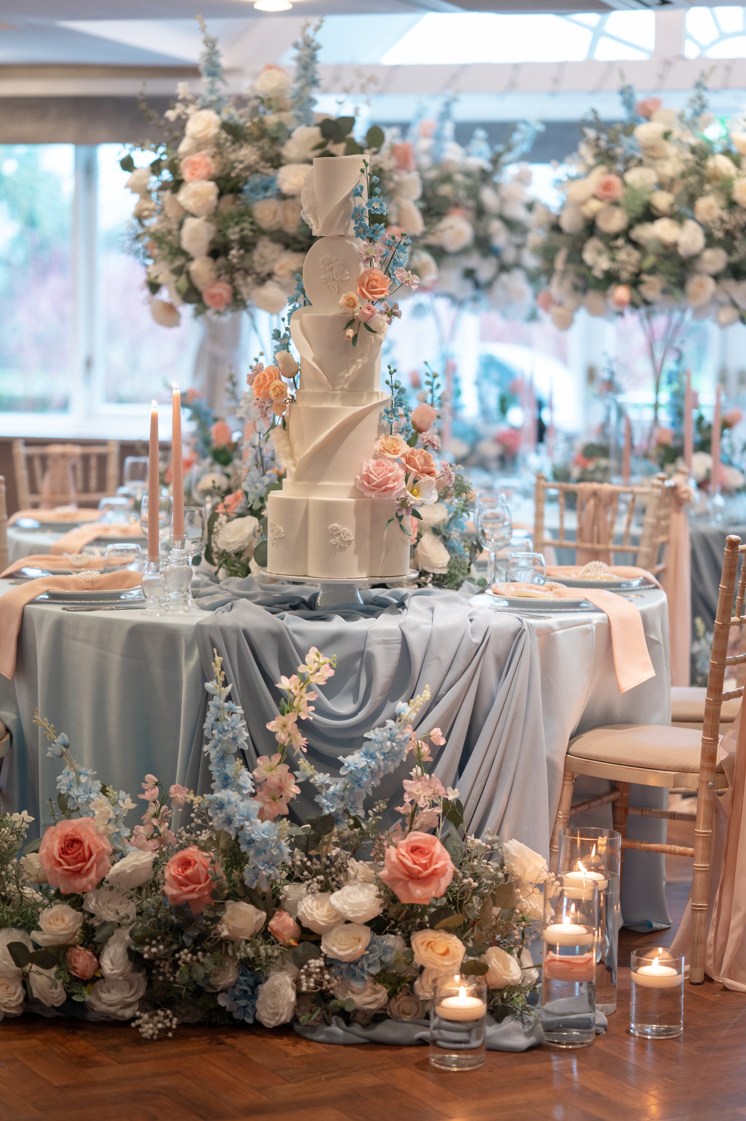 A wedding cake decorated with pastel-colored flowers on a round table, surrounded by candles and a floral arrangement with pink and white roses and blue flowers in a grand hall.