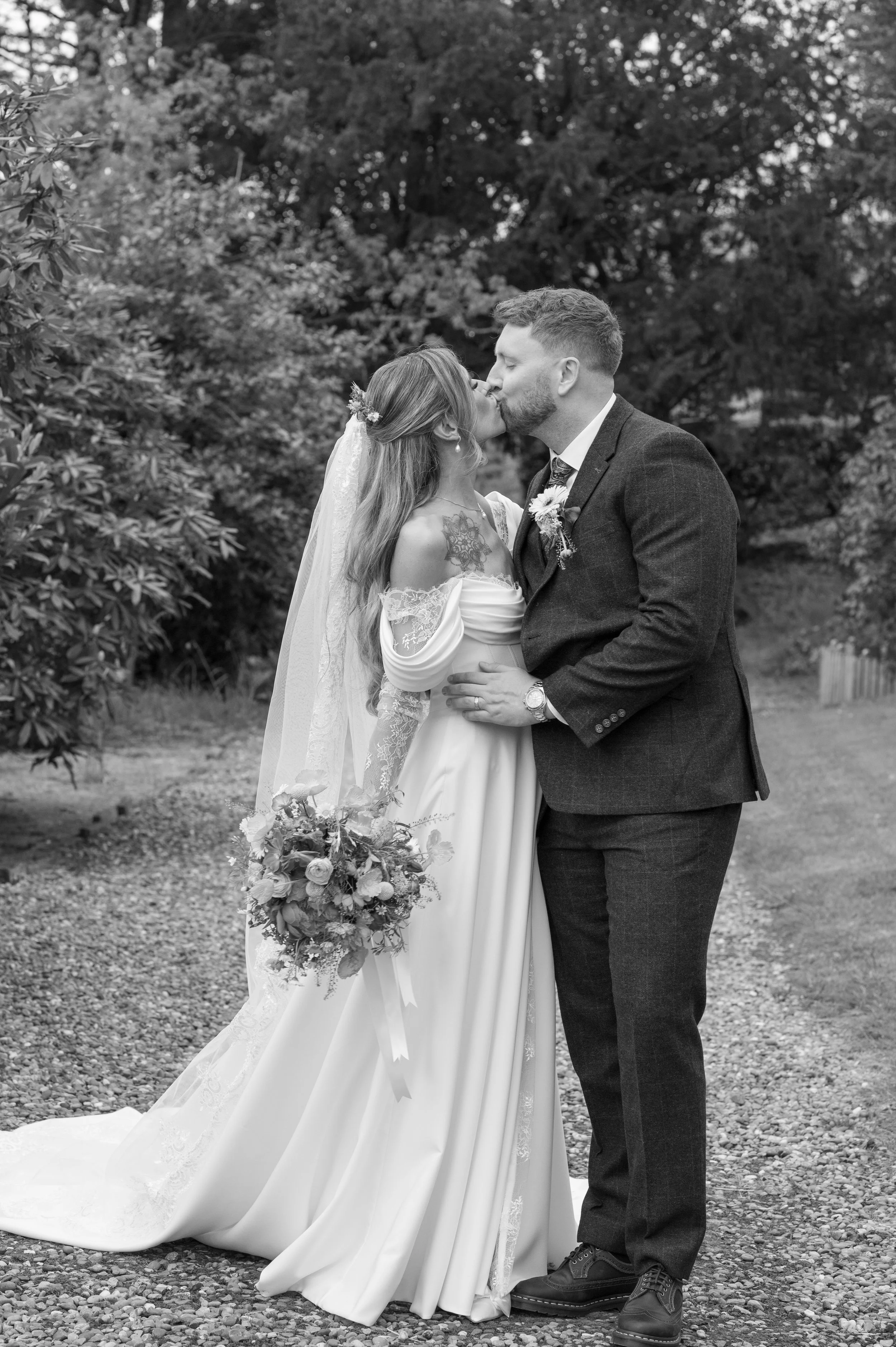 A bride and groom share a kiss outdoors during their wedding, surrounded by trees and natural scenery. The bride is in a wedding dress holding a bouquet, and the groom is in a suit.