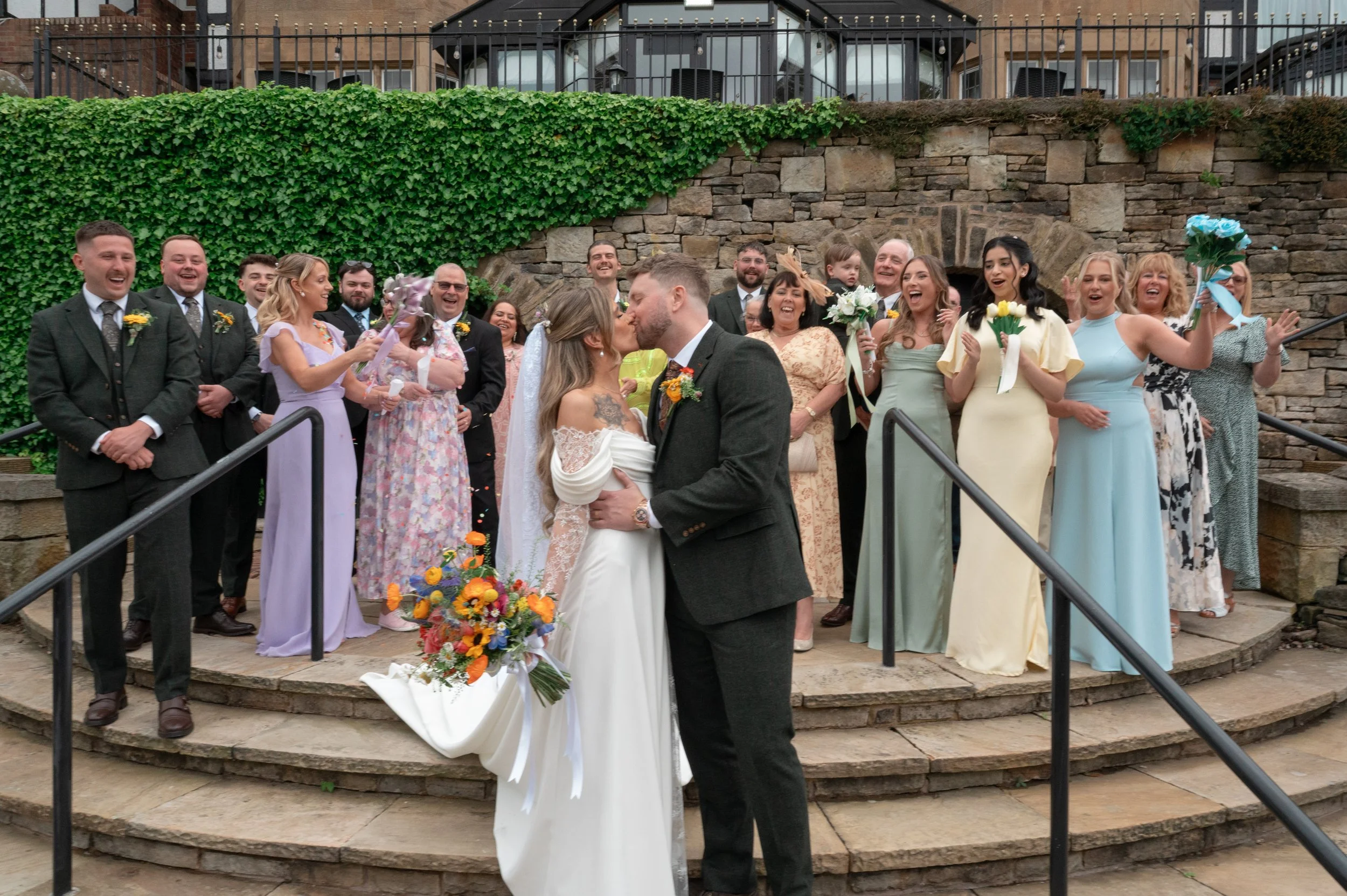A wedding celebration with a bride and groom kissing on steps, surrounded by smiling guests holding flowers, in front of a stone wall and greenery.