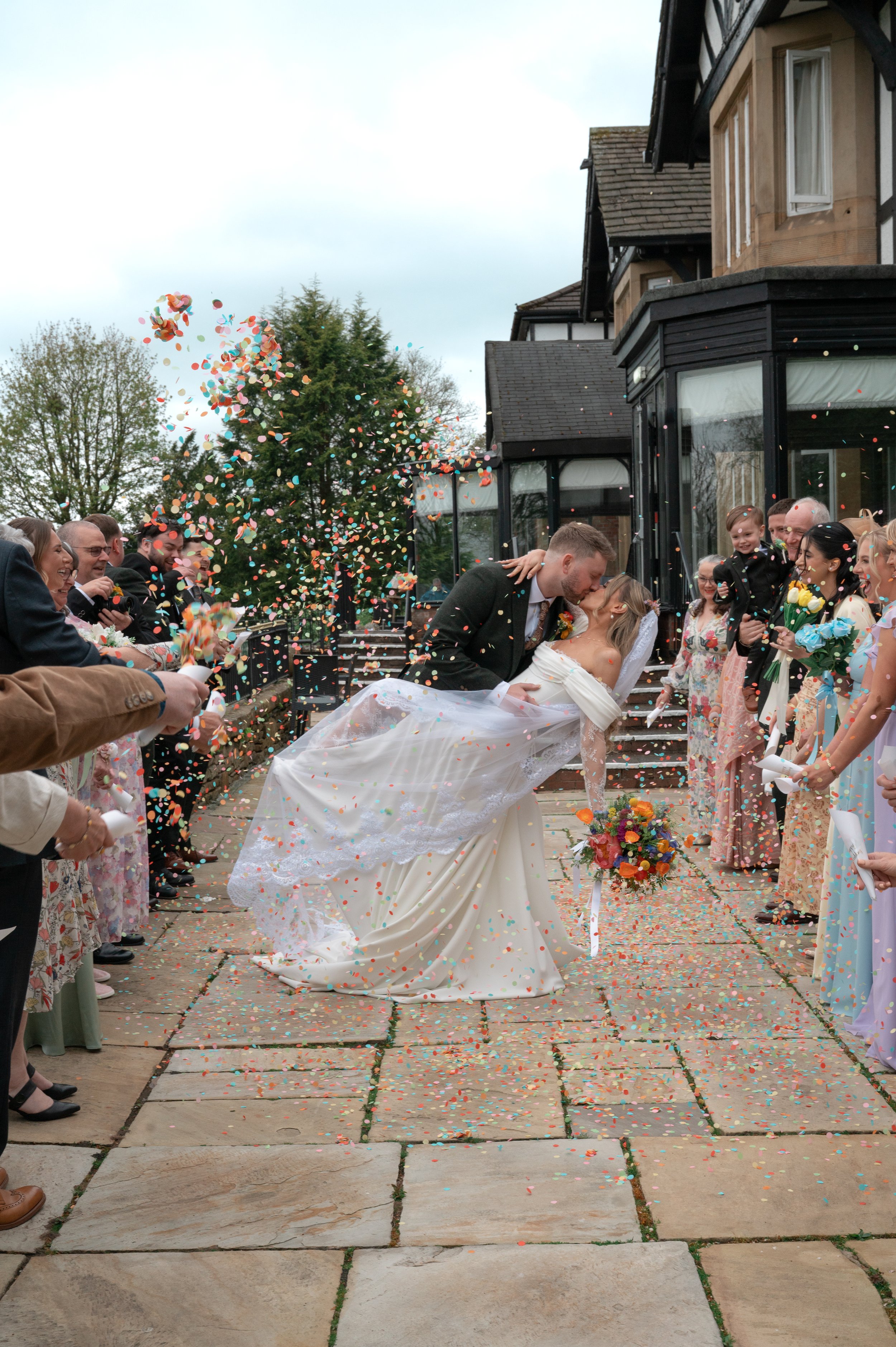A bride and groom kiss while surrounded by friends and family outside a house, with colorful confetti falling around them after their wedding ceremony.