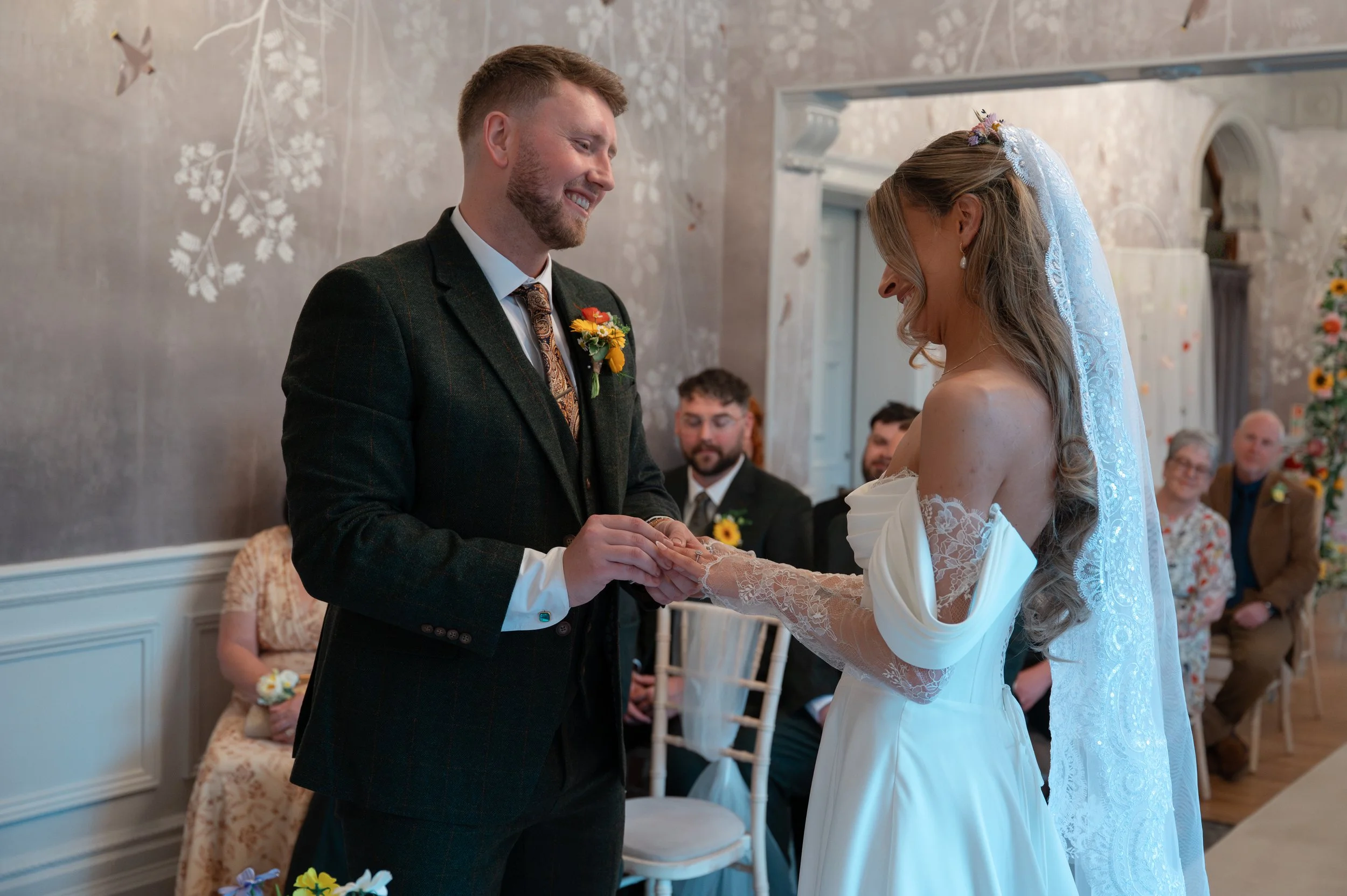 A bride and groom exchanging vows during a wedding ceremony indoors. The groom is wearing a dark suit with a patterned tie, and the bride is dressed in a white gown with lace sleeves and a veil. Guests are seated in the background, and the room has f