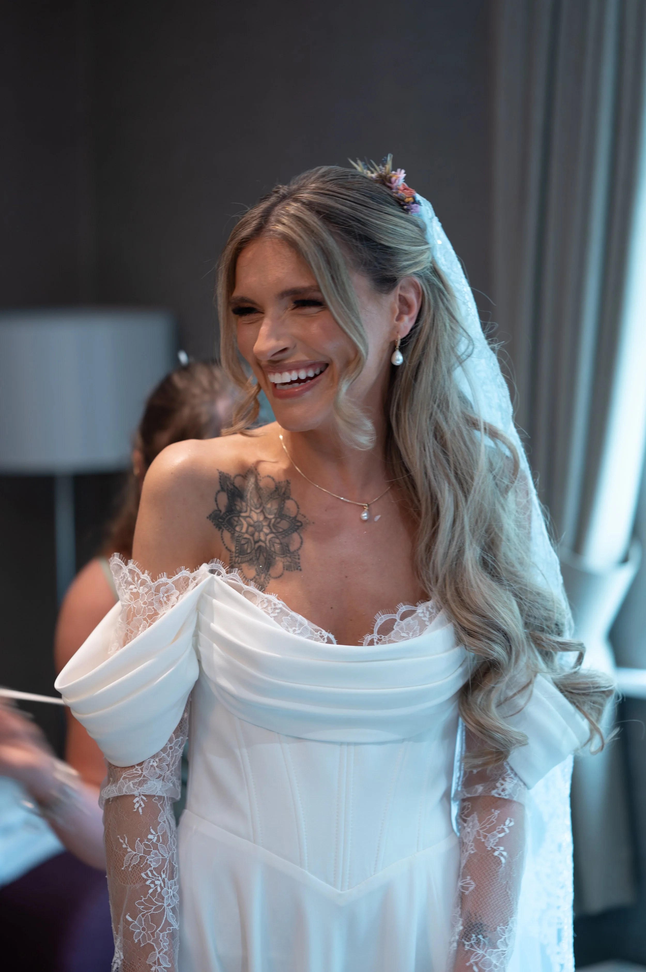 A woman in a white wedding dress with lace sleeves and a floral hair accessory, smiling and looking to the side in a softly lit room.